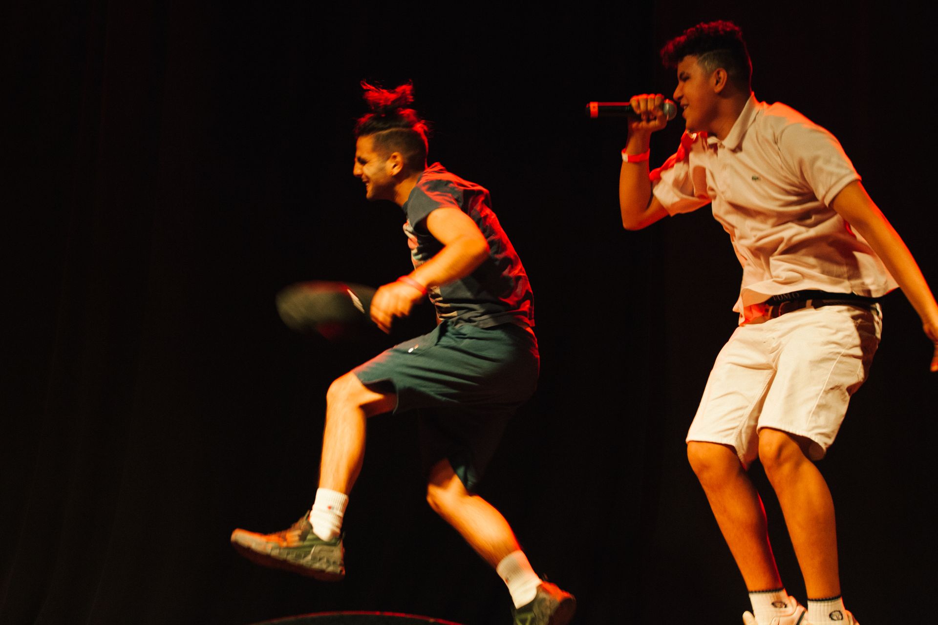 Due uomini sul palco, uno salta con un pallone da basket, l'altro tiene un microfono, mentre si esibiscono. Sfondo scuro.