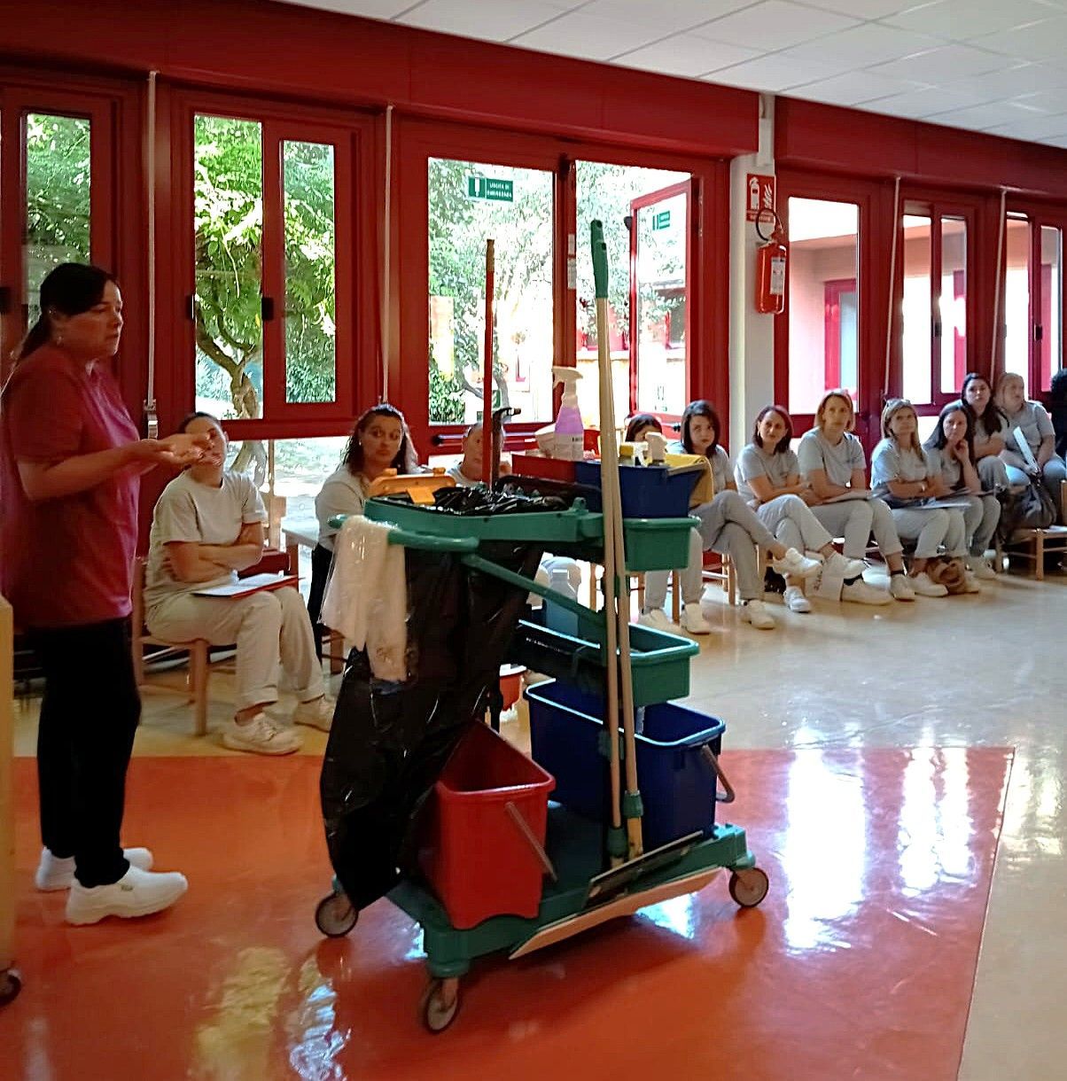 Una donna mostra gli strumenti per la pulizia a un gruppo di persone sedute in una stanza ben illuminata.