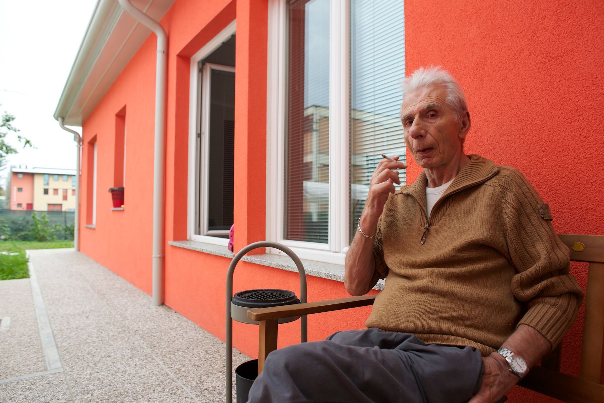 Un uomo anziano è seduto su una panchina fuori da un edificio color corallo e fuma.