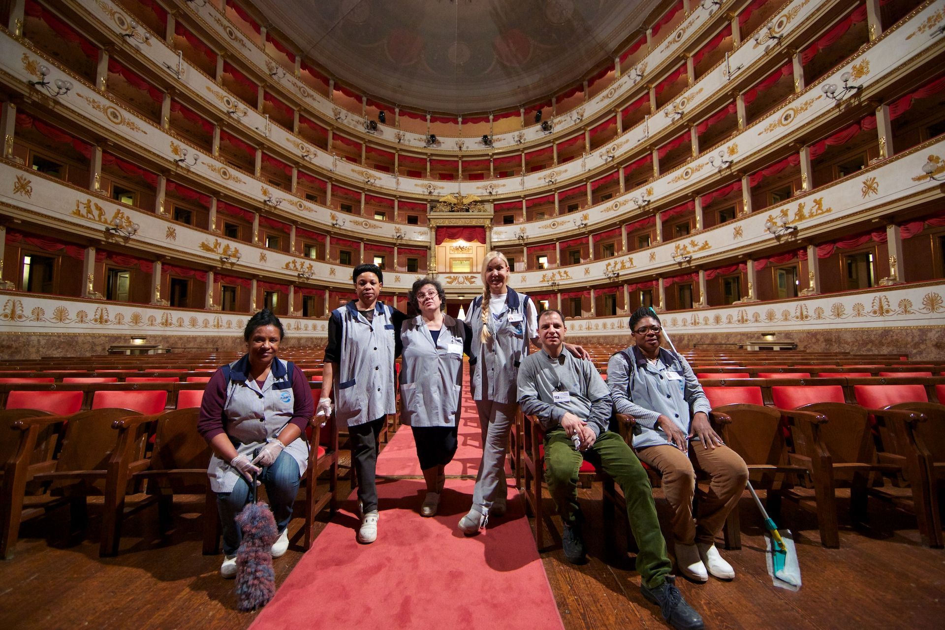 Un gruppo di sei persone in posa in un teatro vuoto. Indossano uniformi da lavoro coordinate e sorridono alla telecamera. Sedili rossi e decorazioni dorate.