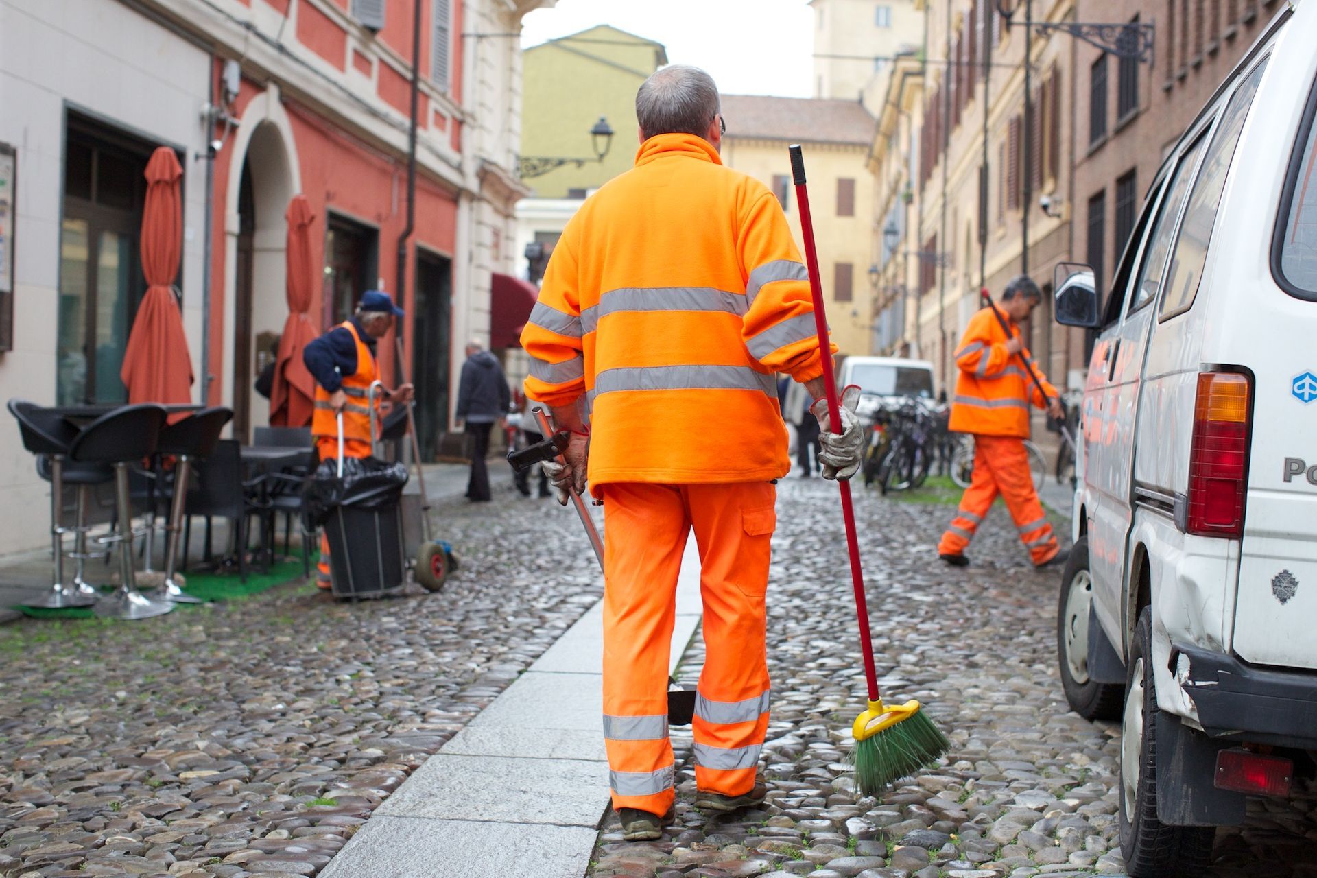 Gli spazzini con i giubbotti arancioni spazzano una strada acciottolata vicino a un'auto parcheggiata.