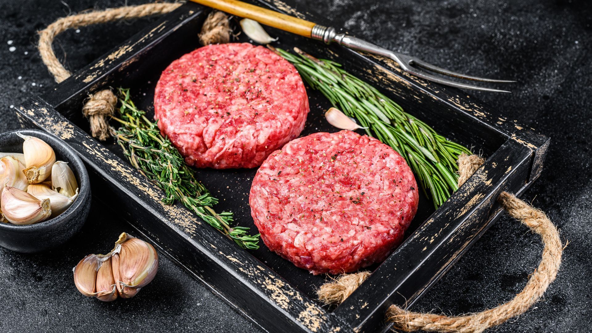 to rå hamburgerfrikadeller i en træbakke med hvidløg og rosmarin
