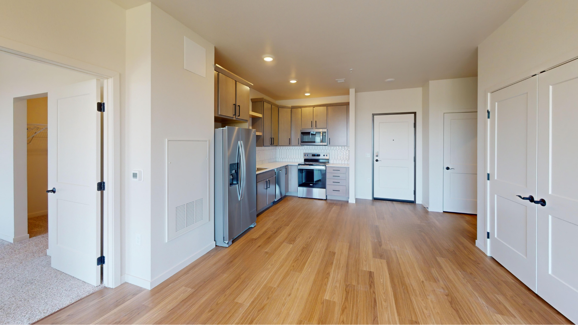 Open-concept apartment interior with kitchen and living space. Features wood floors, stainless steel appliances, and neutral-toned walls.