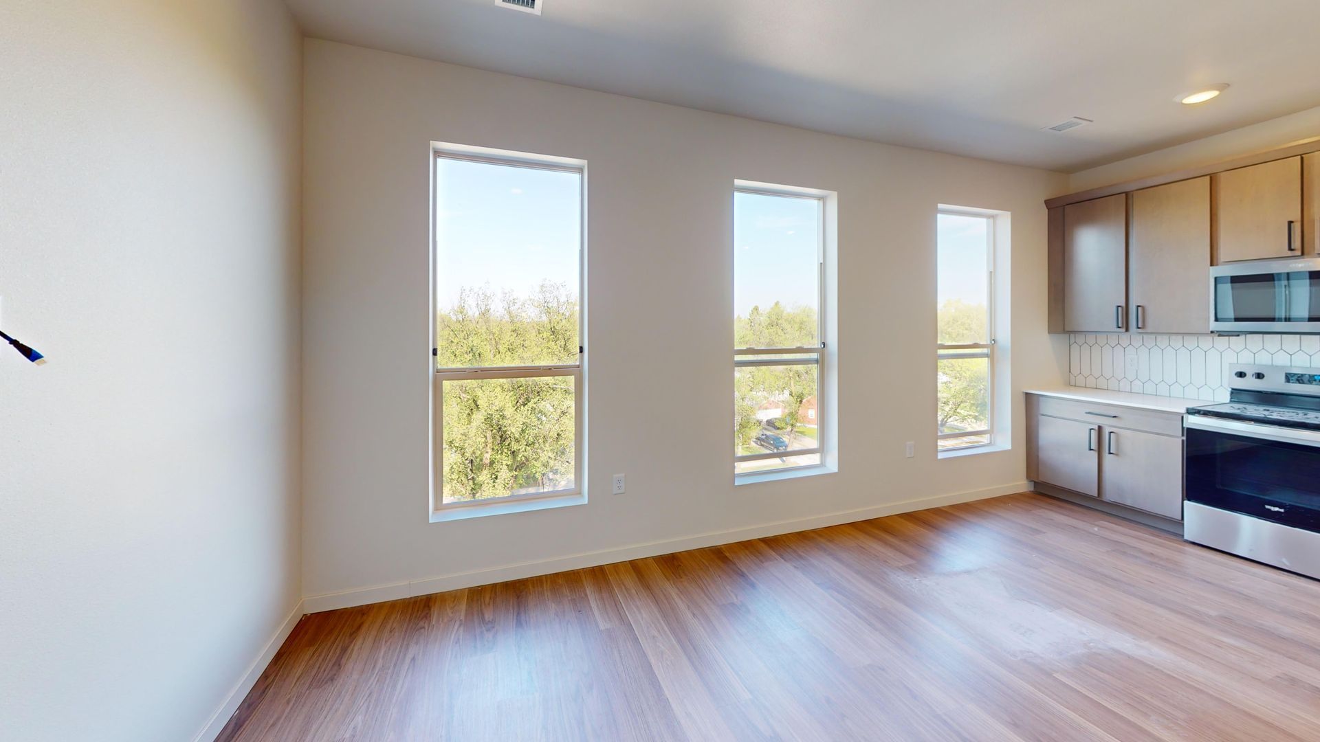 An empty apartment interior with three vertical windows overlooking trees. The kitchen area has light wood cabinets and a stainless steel range.