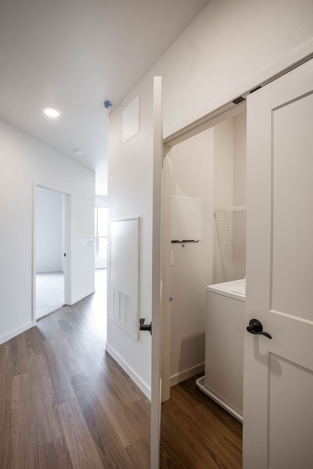 A hallway with wooden flooring, a partially open white door reveals a laundry area with a washing machine.