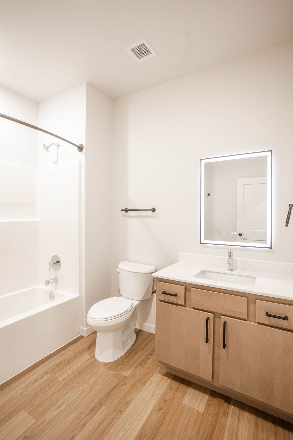 A modern bathroom with a white tub, toilet, and sink with a lighted mirror. The floor is light wood.