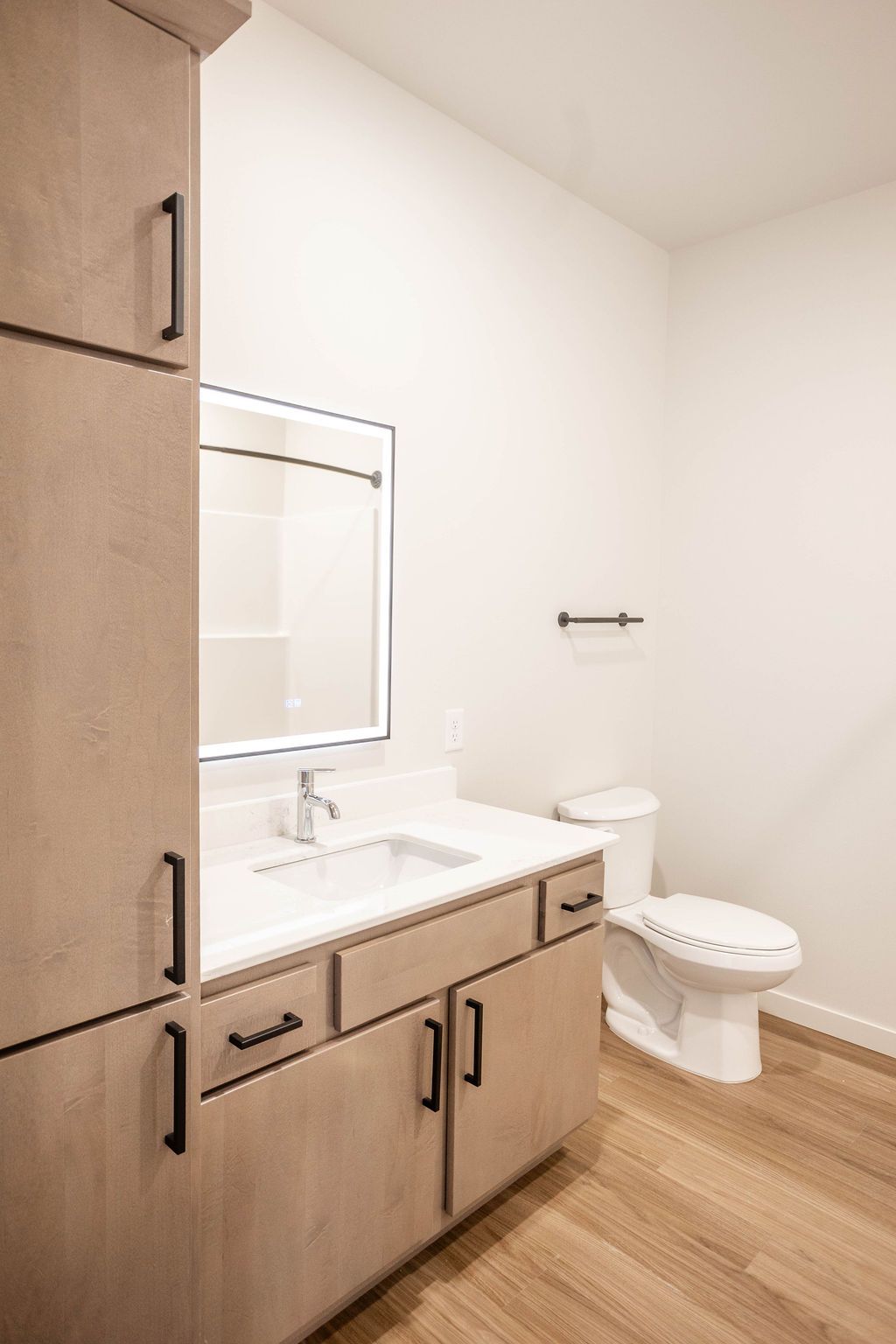 Modern bathroom with light wood cabinets and flooring, a white sink and toilet, and a large mirror with a lit frame.