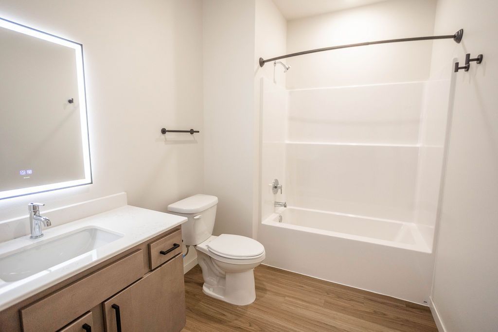 Modern bathroom with a vanity, toilet, and bathtub/shower combo; beige cabinets, light-colored walls, and wood-look flooring.