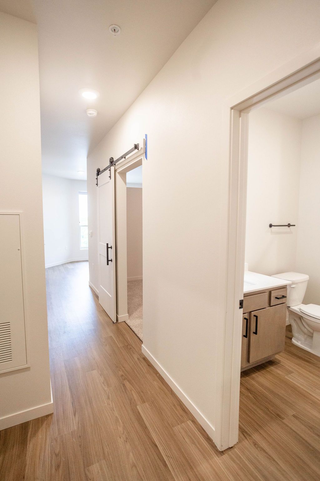 Interior hallway with wood flooring, white walls, and a bathroom visible on the right. A sliding barn door is in the center.