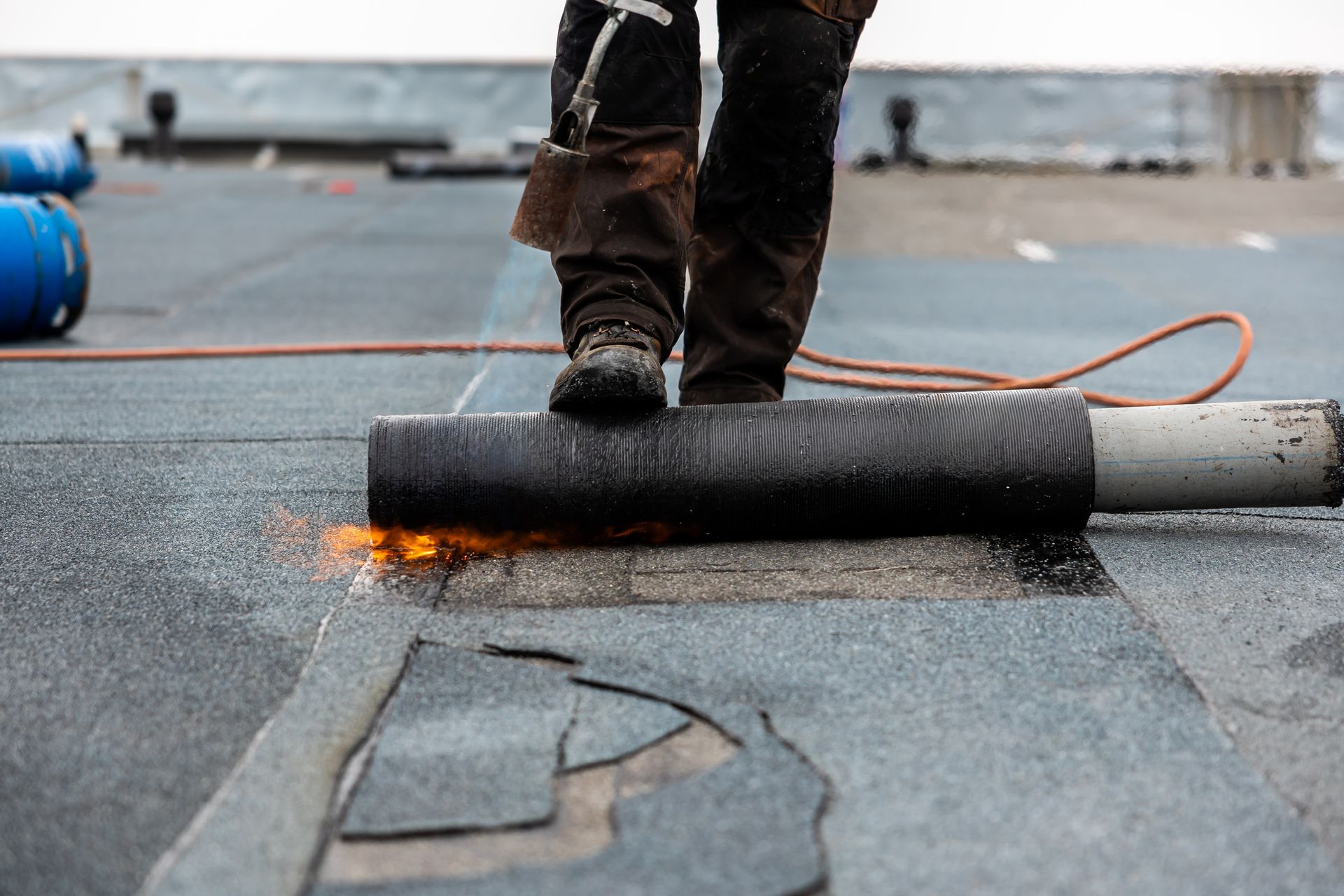 Roofer torching a rolled roofing membrane onto a dark surface, with flames visible. Person in boots standing nearby.
