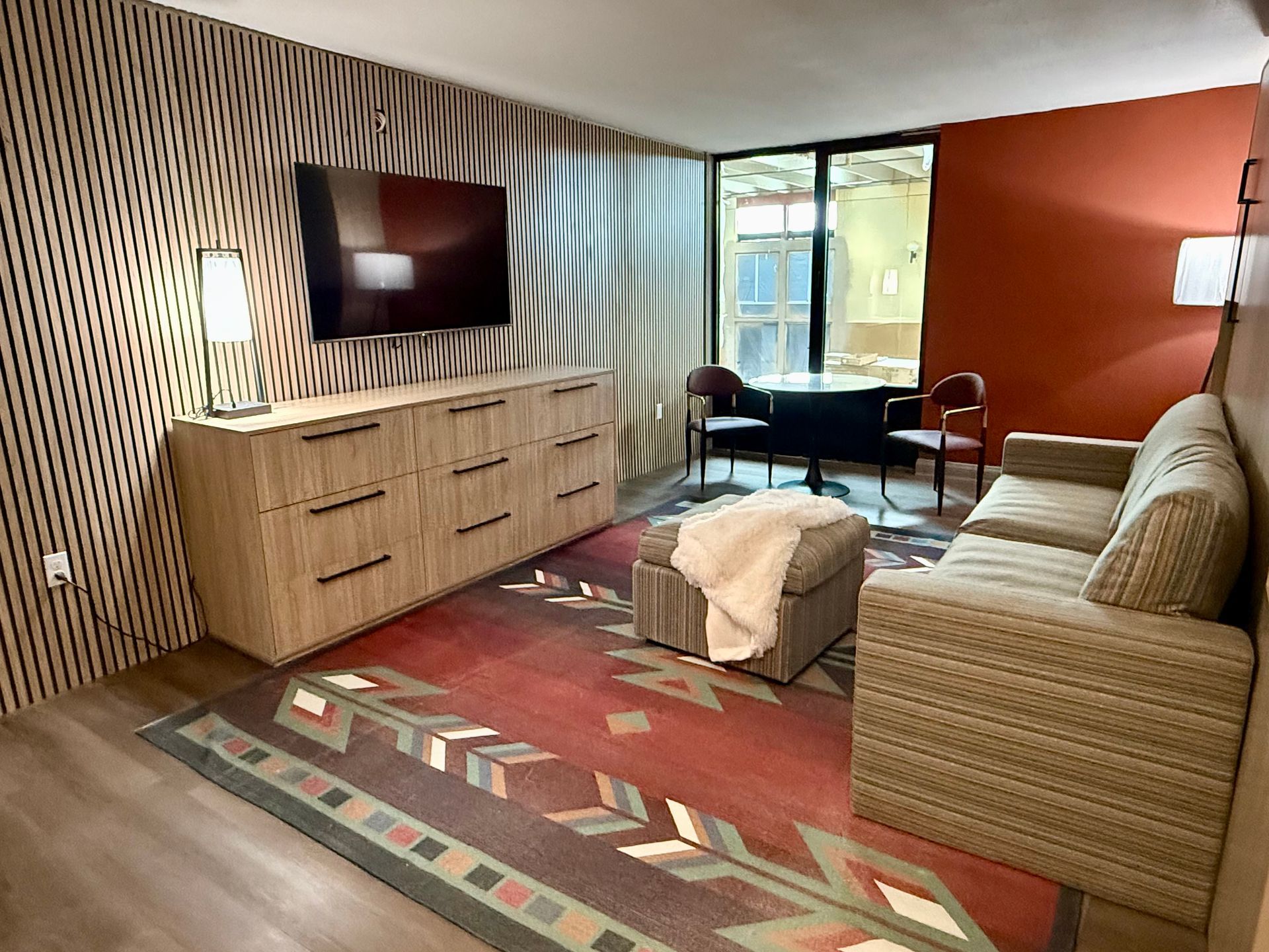 Photo showing a living room with a rug, couch, chest of drawers, television, and a table with two chairs next to a window