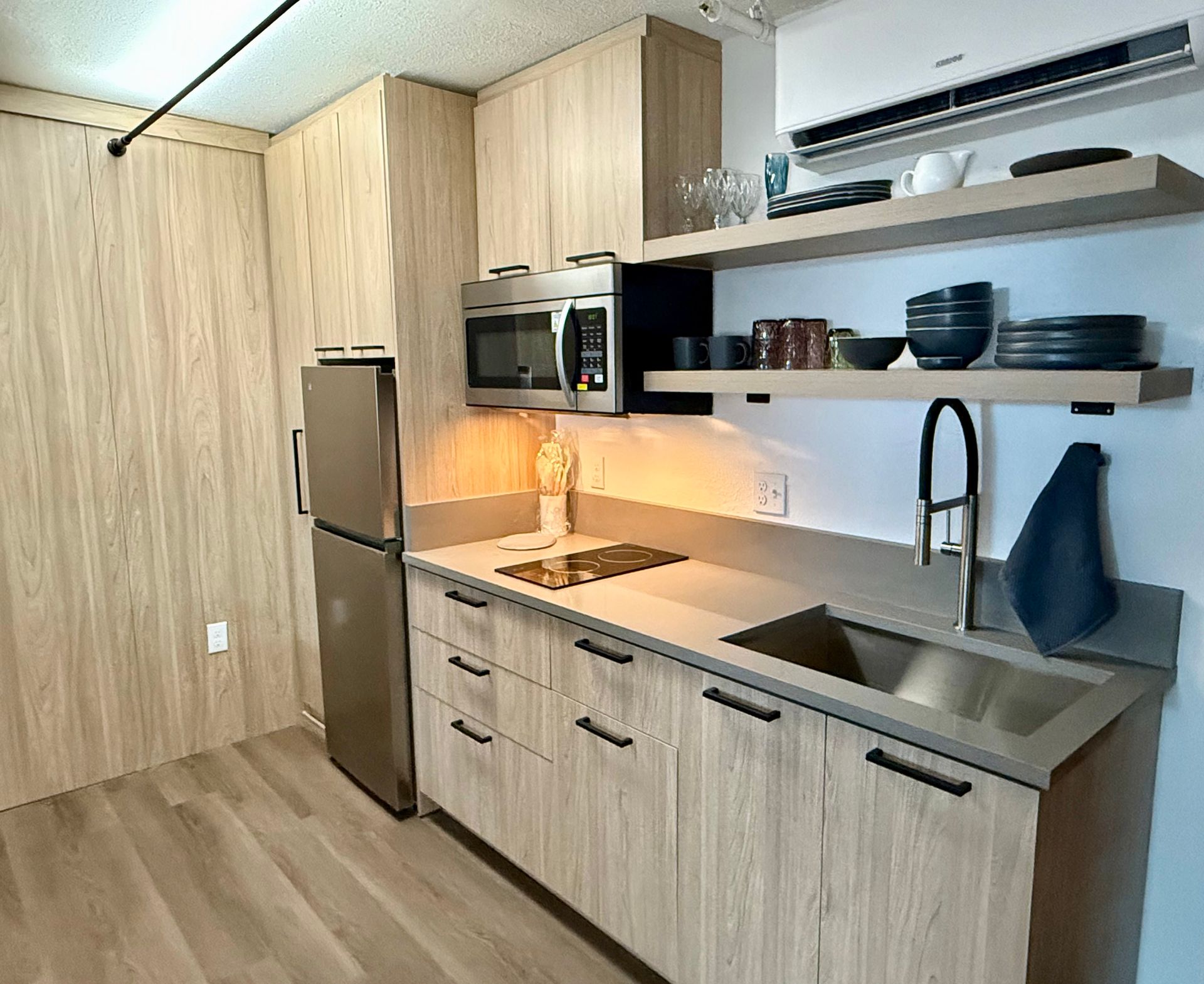 Photo showing a refrigerator tucked into a wall, a large sink, microwave, dishes and shelving