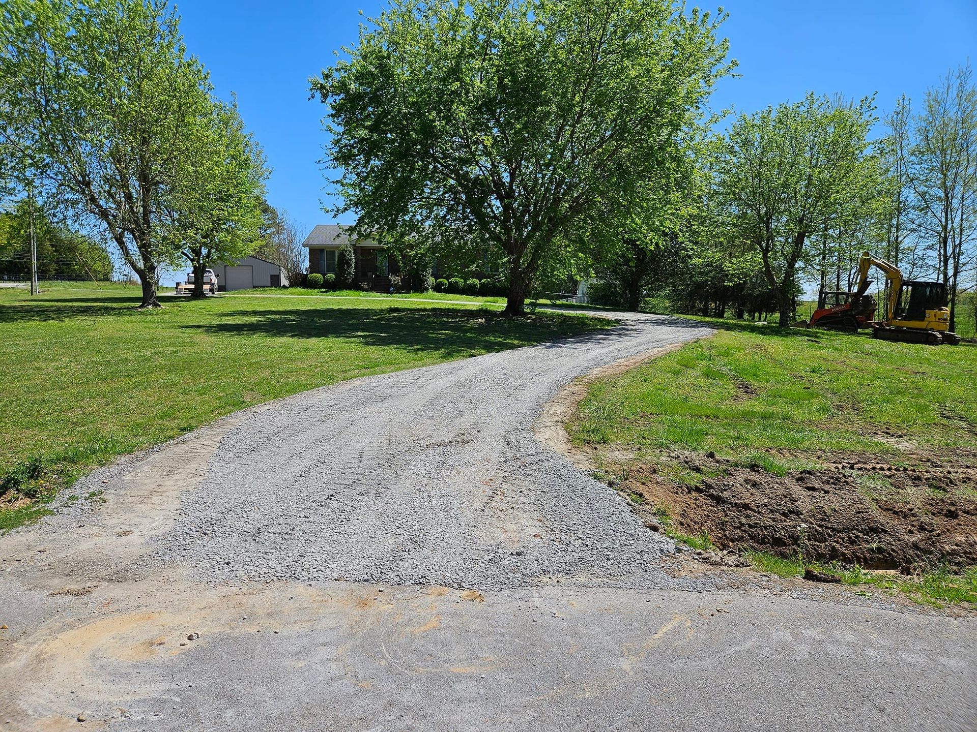 Gravel driveway leading to a house, flanked by green grass and trees under a clear blue sky.
