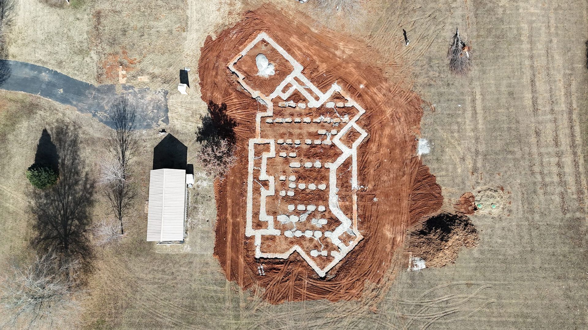 Overhead view of excavated building foundations in a brown field. A small building with a light roof is to the left.