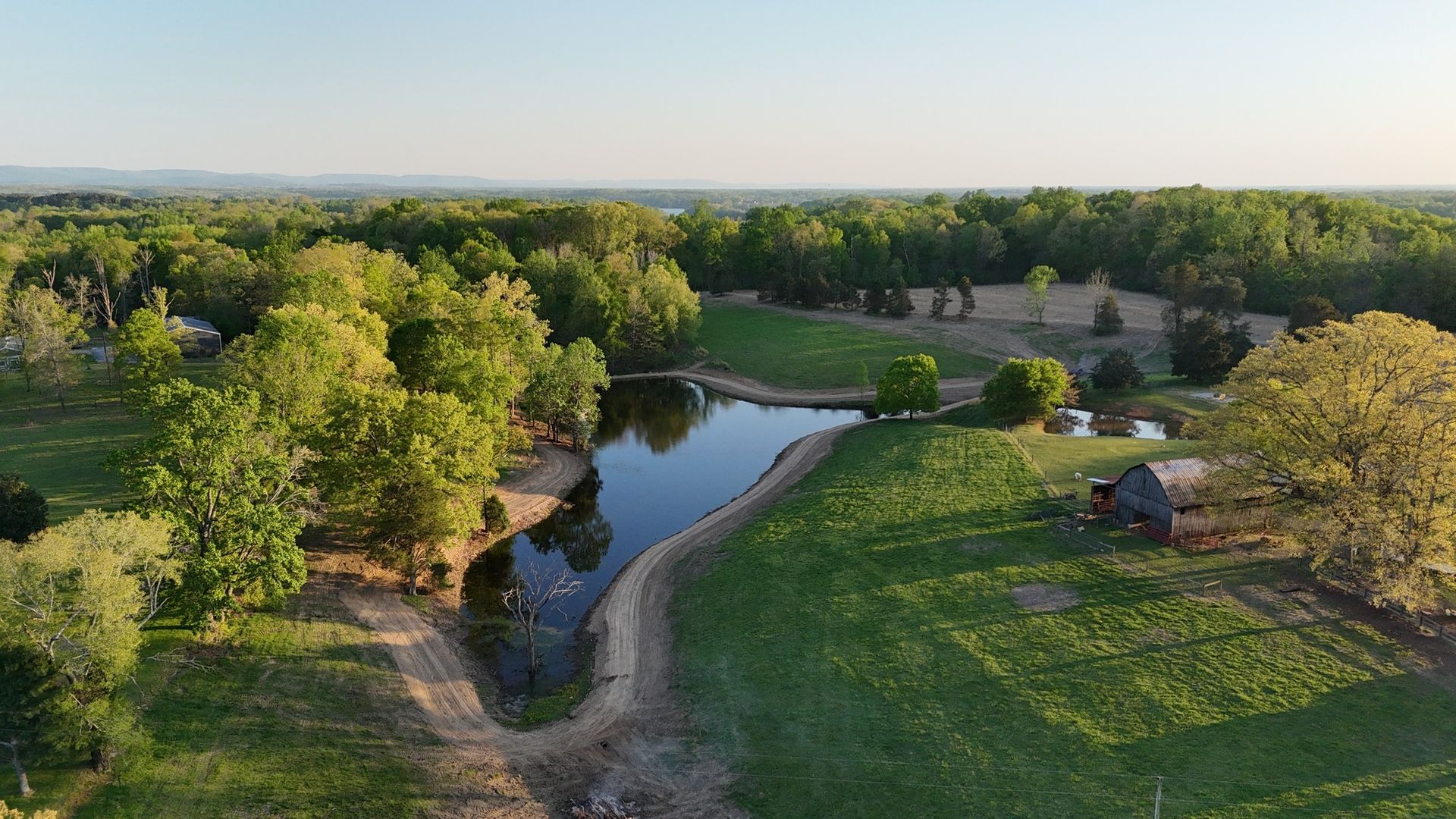 Lush green landscape with pond reflecting trees, surrounded by grassy fields and a small wooden structure under a clear sky.