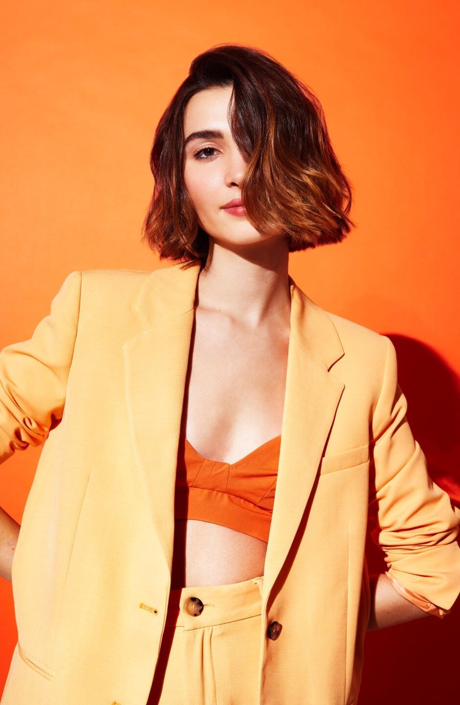 Donna con capelli biondi corti e ondulati, che indossa un top bianco trasparente con dettagli elaborati, in posa, sfondo nero.