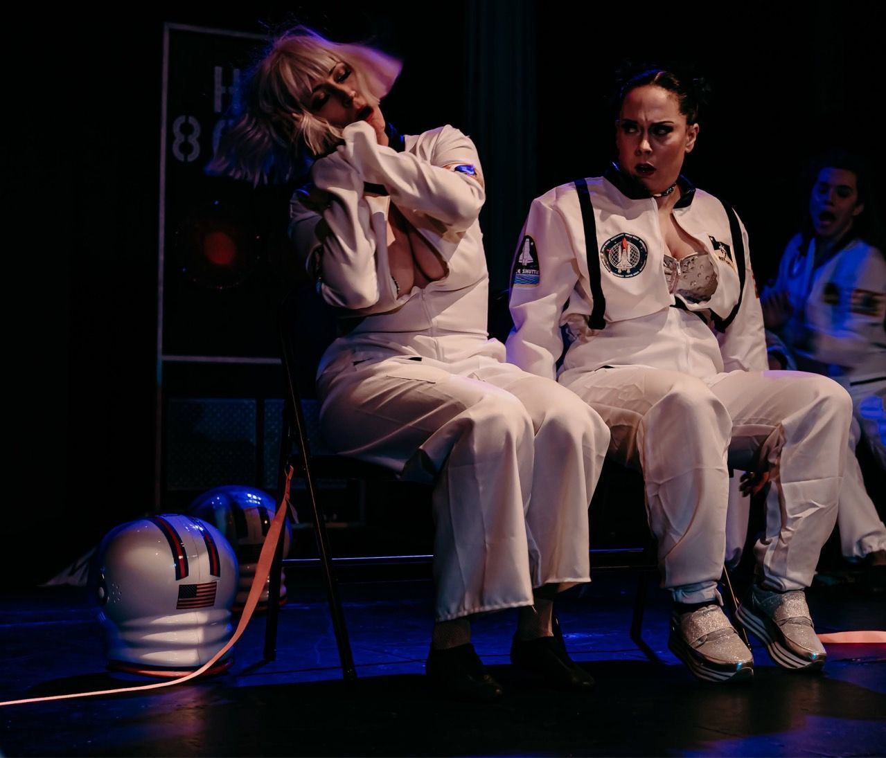 Two astronauts are sitting on a stage with a helmet in the background
