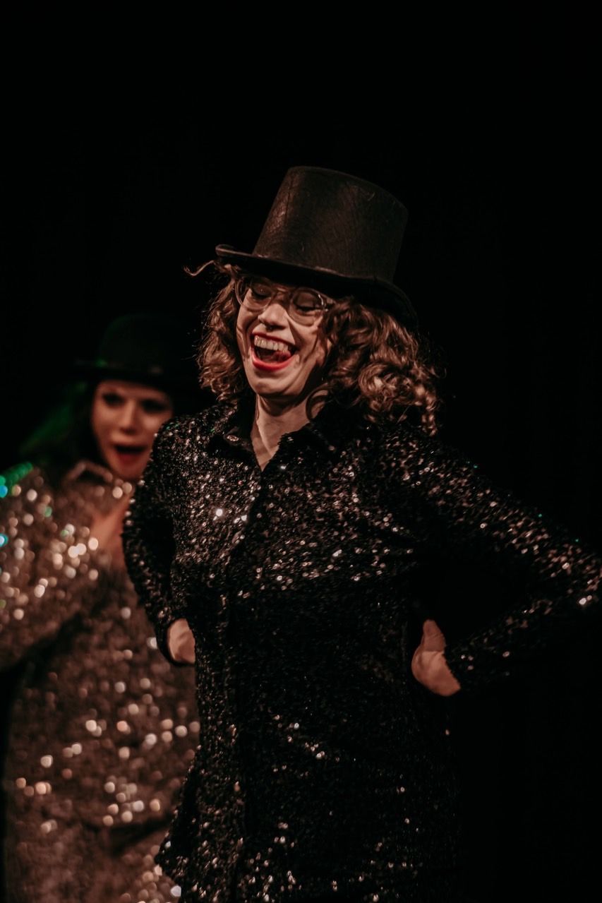 A woman in a top hat and sequined shirt is dancing on a stage.