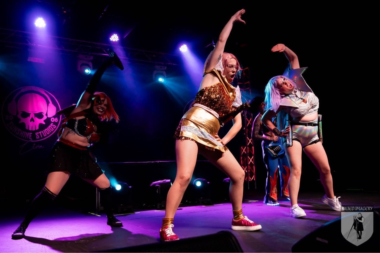 A group of women are dancing on a stage with a skull in the background.