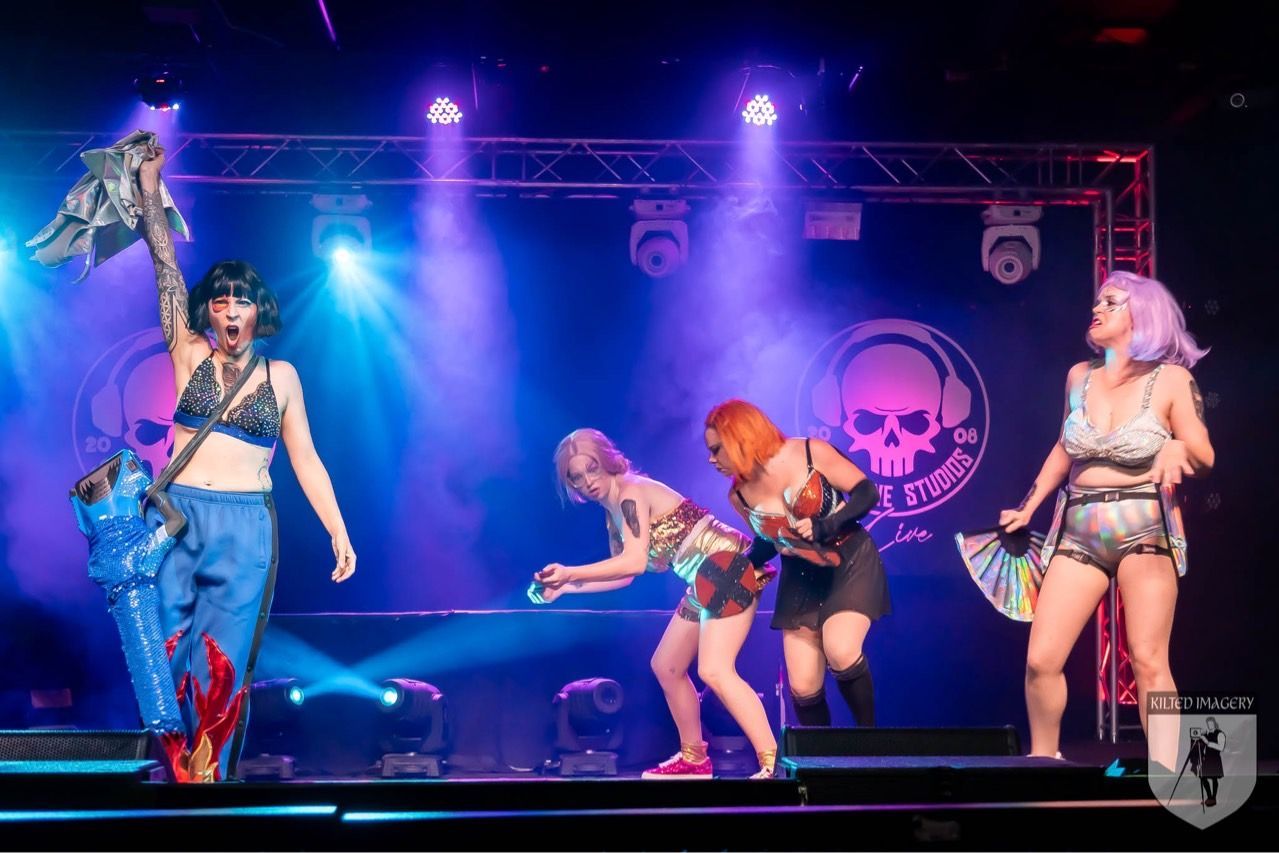 A group of women are dancing on a stage in front of a skull logo.