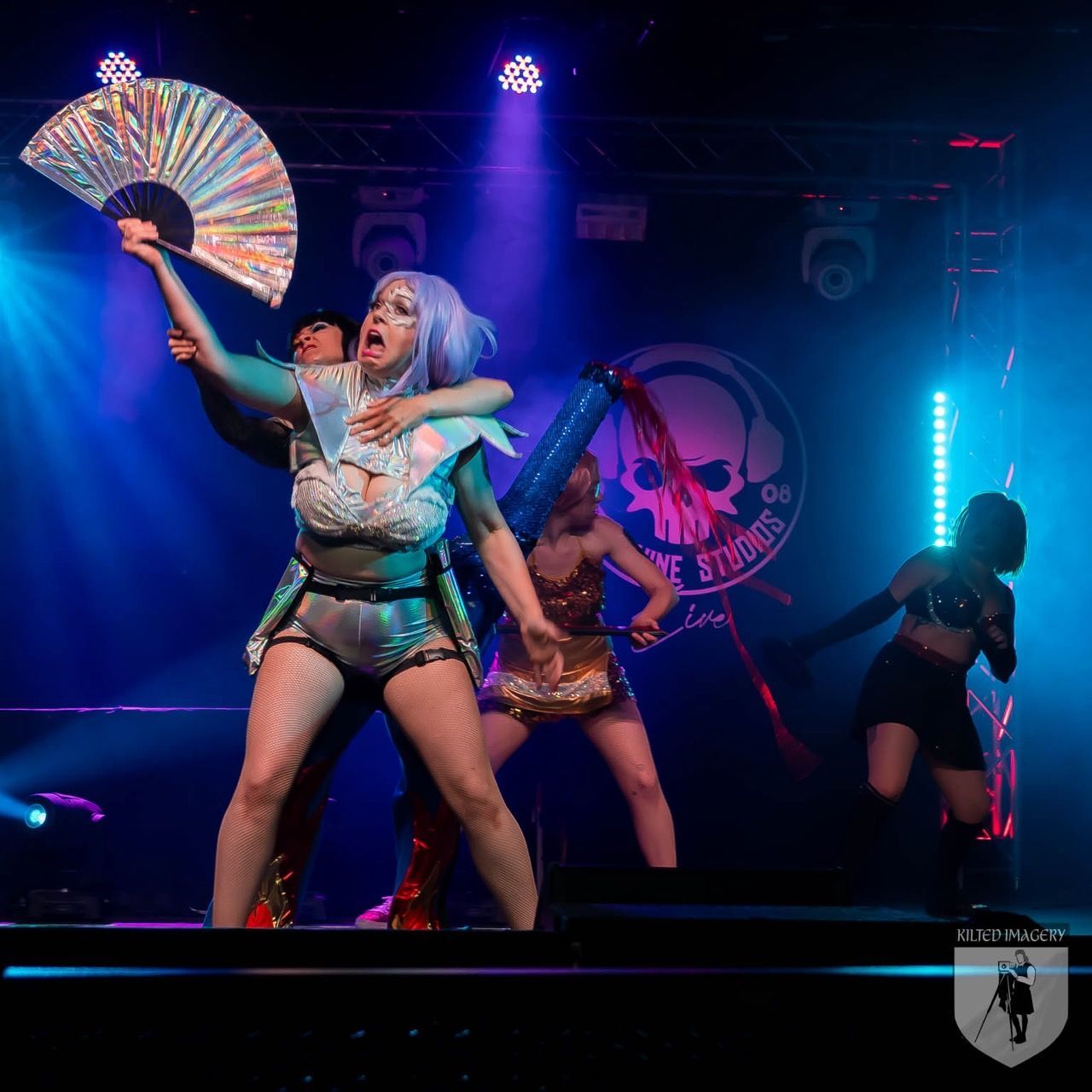 A woman holding a fan on a stage with a skull logo in the background
