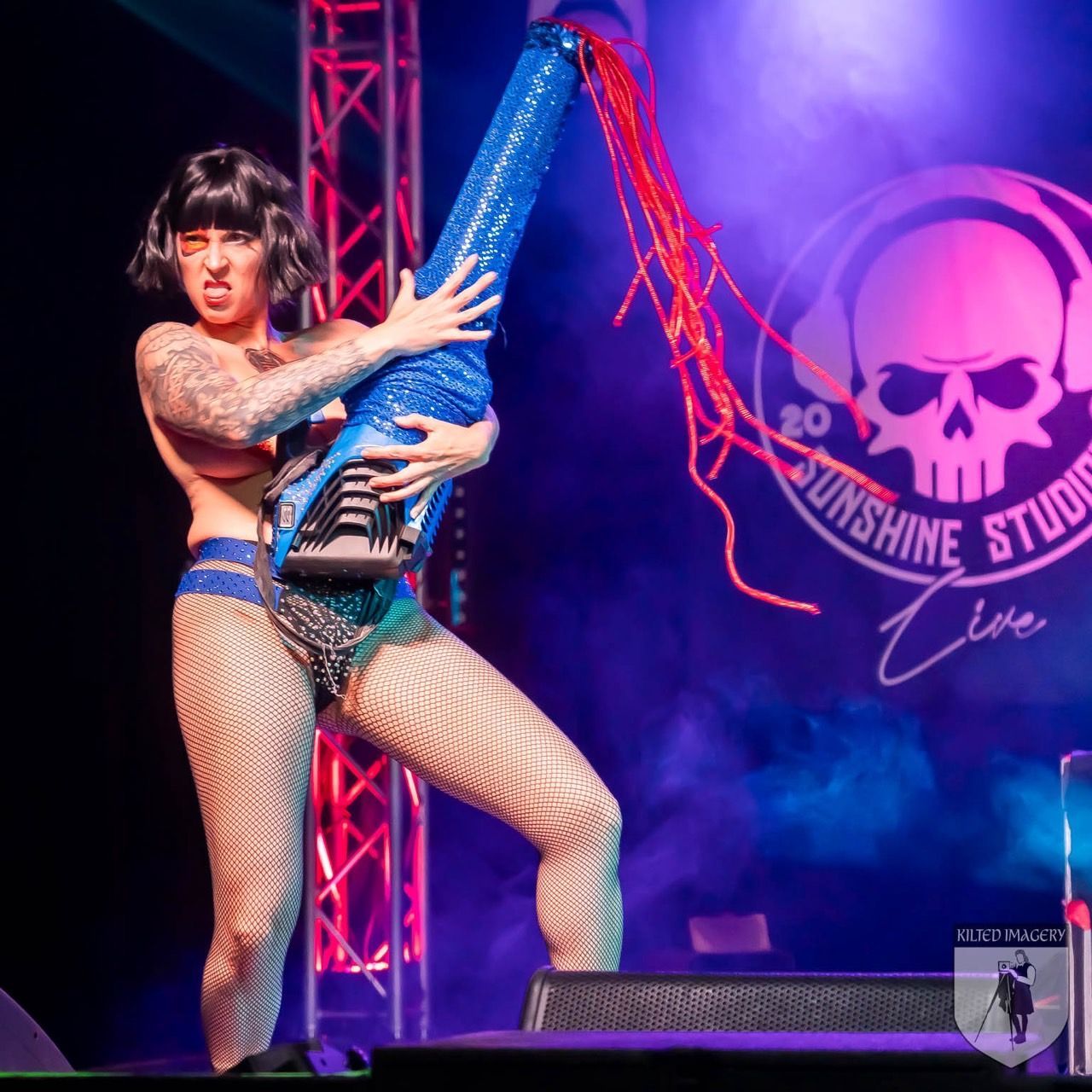 A woman in fishnet stockings is holding a guitar in front of a sign that says sunshine studio live