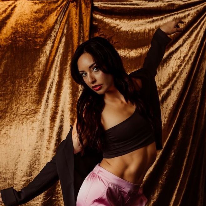 A woman in a black crop top and pink shorts is standing in front of a gold curtain