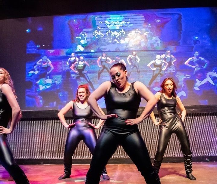 A group of women are dancing on a stage in front of a large screen.
