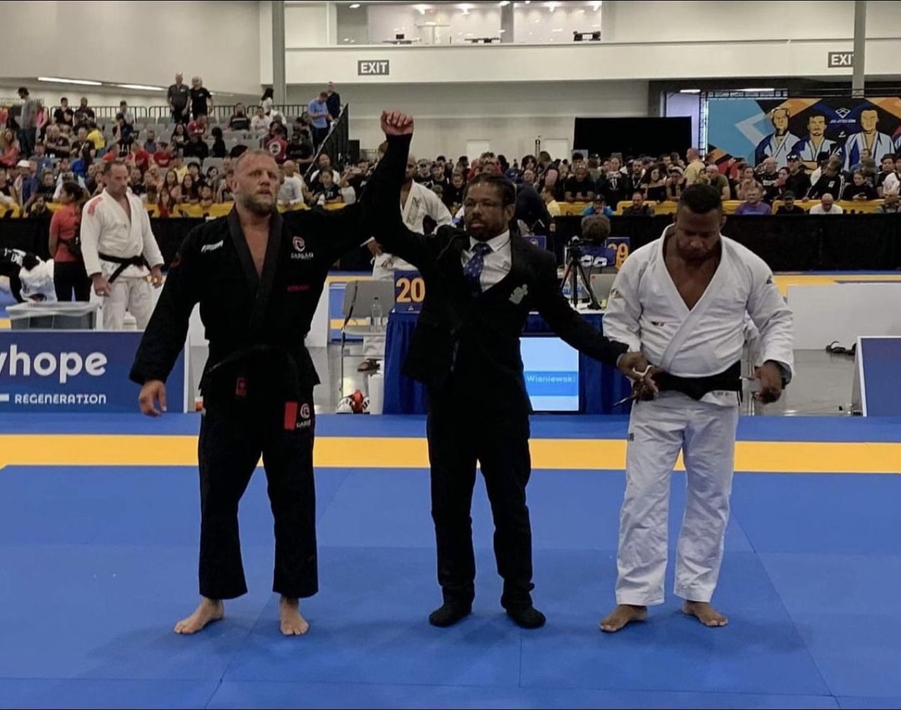 Three men are standing on a wrestling mat and one of them (Christopoher Wisniewski) has his hand up in the air after winning a Jiu Jitsu match.