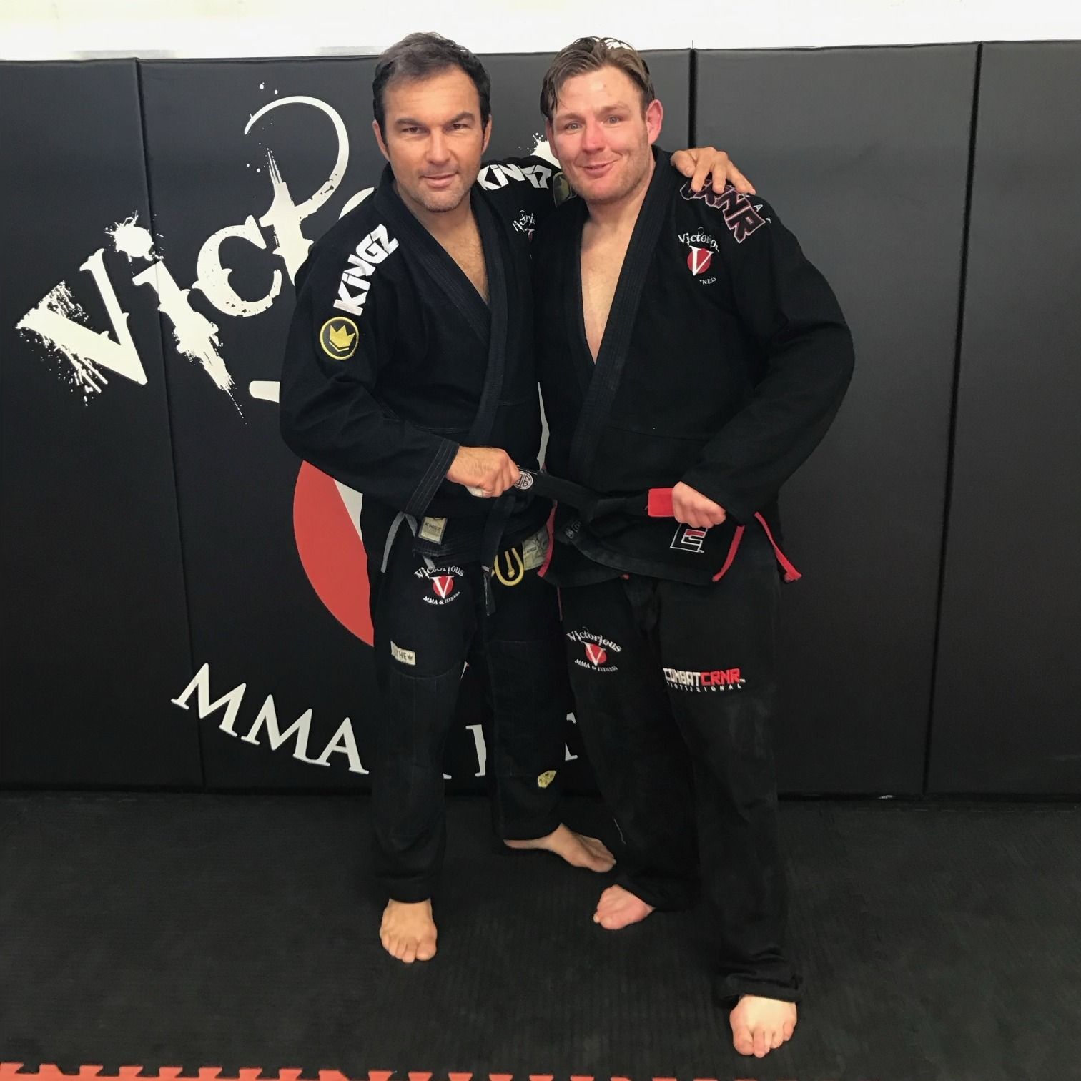 Two men are posing for a picture in front of a wall that says Victorious MMA.