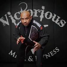 Victor Torres is wearing a black Jiu Jitsu gi standing in front of a black wall.