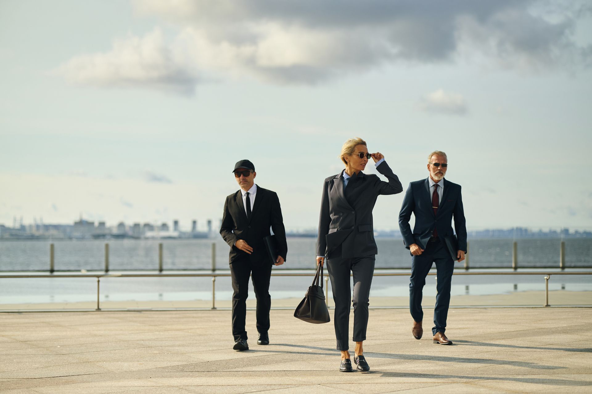Three people in suits walking on a paved area near water, two men flank a woman, sunny day.
