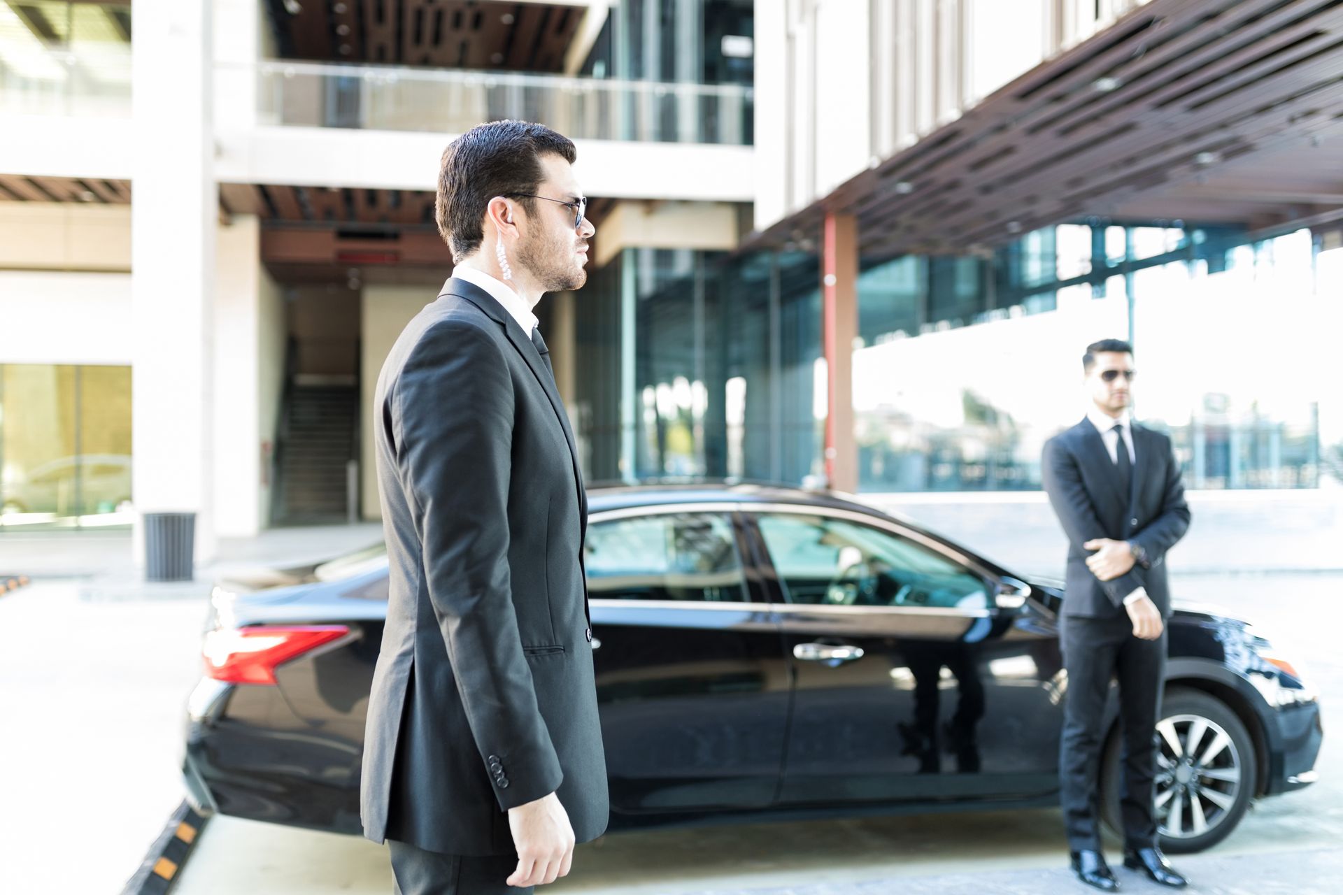 Two men in suits and sunglasses stand by a black car outside a modern building.
