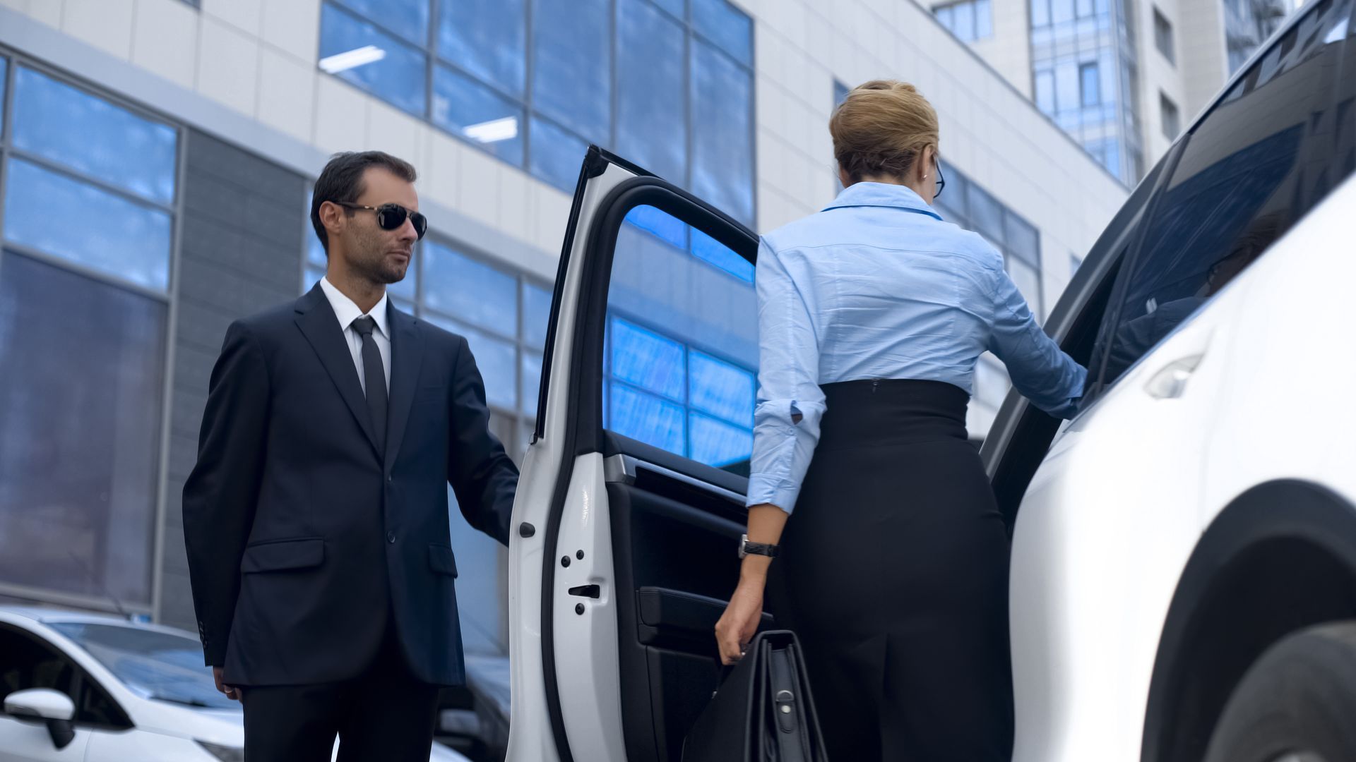 Man in suit opens car door for woman in business attire. Outside a building.