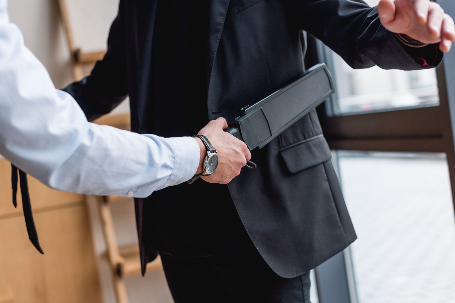 Security guard using a handheld metal detector on a person's jacket in a building.