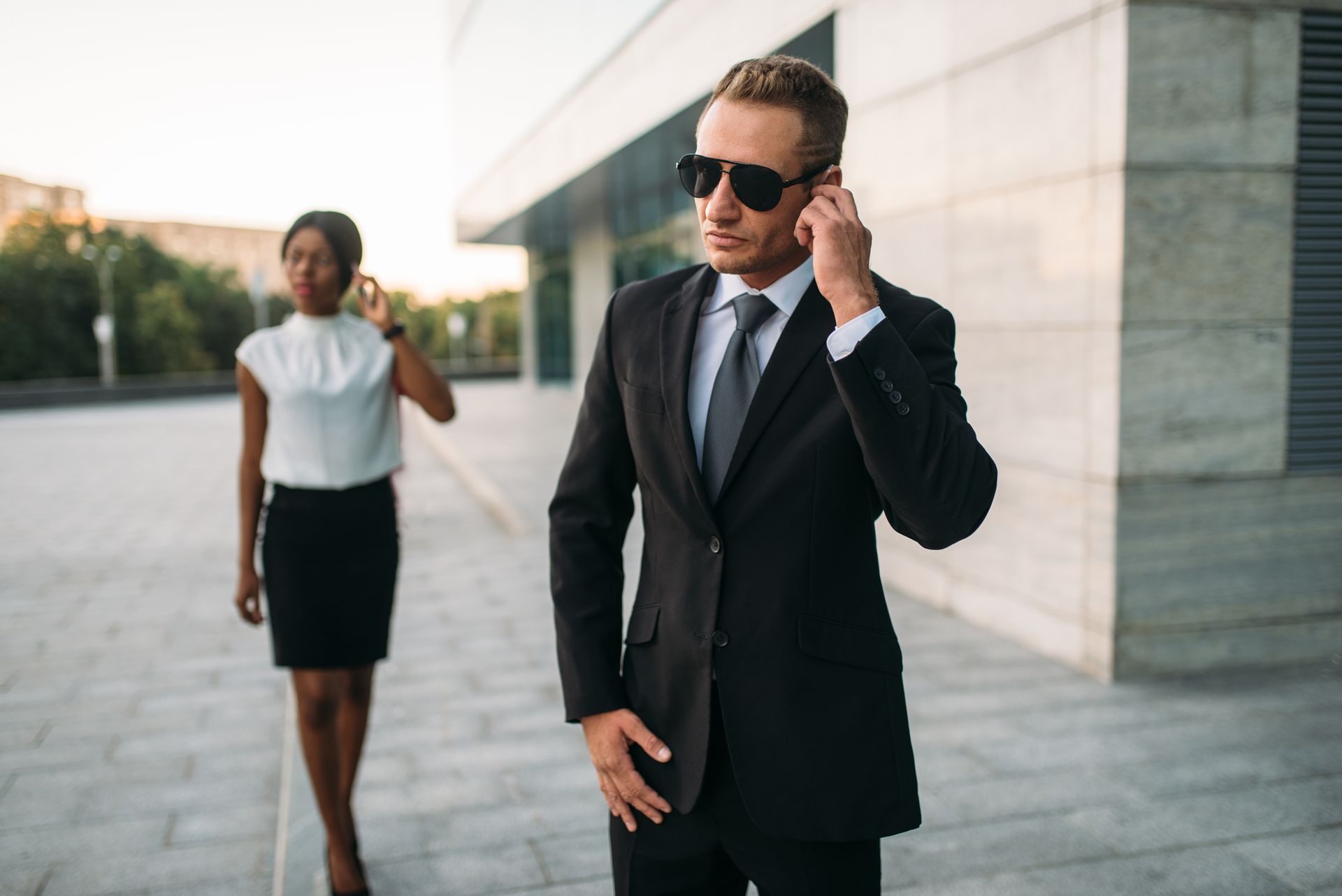 Two people in business attire, possibly security, outside a building, using earpieces.