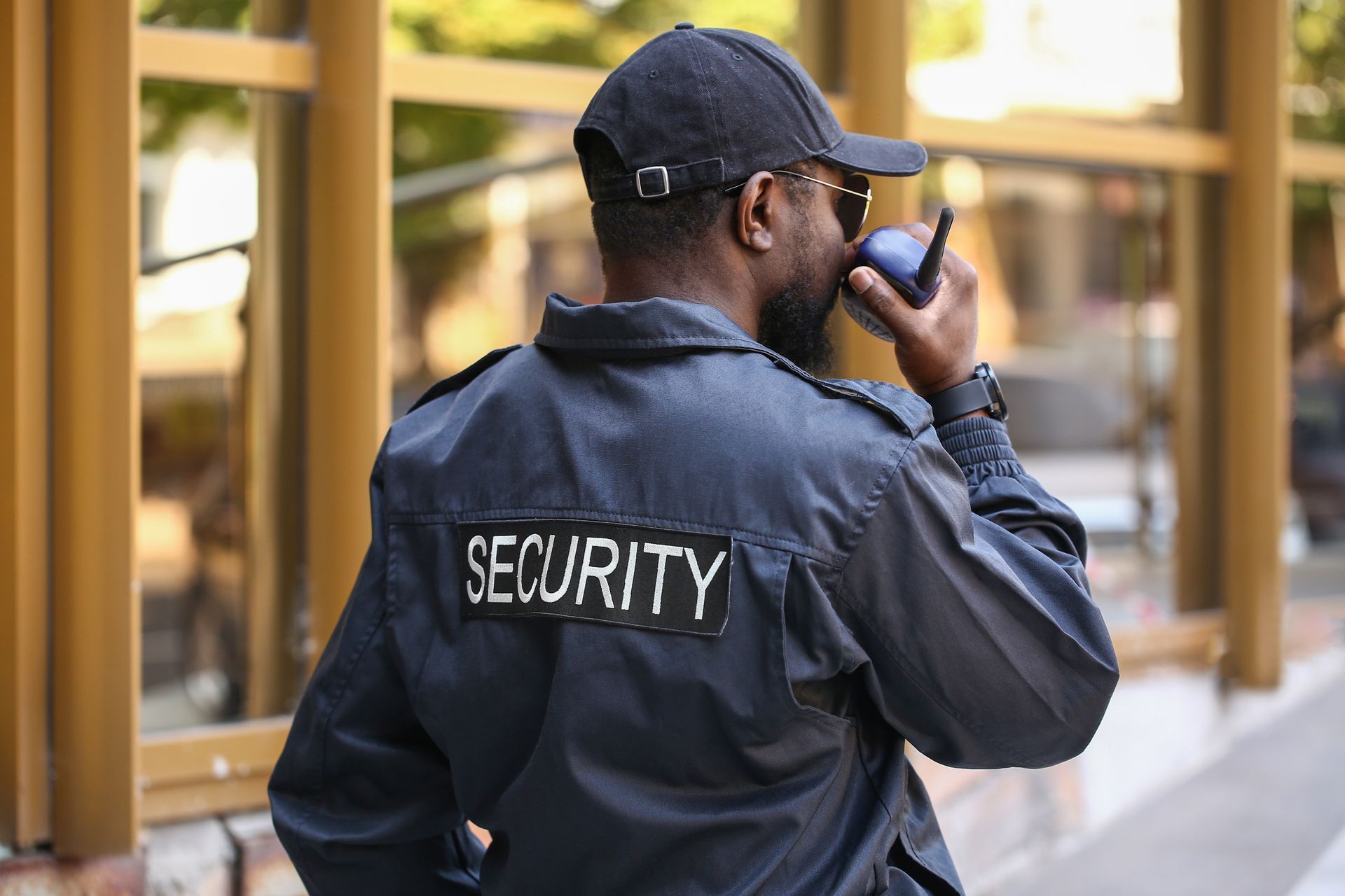 Security guard in a black uniform, cap, and sunglasses, using a walkie-talkie outside a building.
