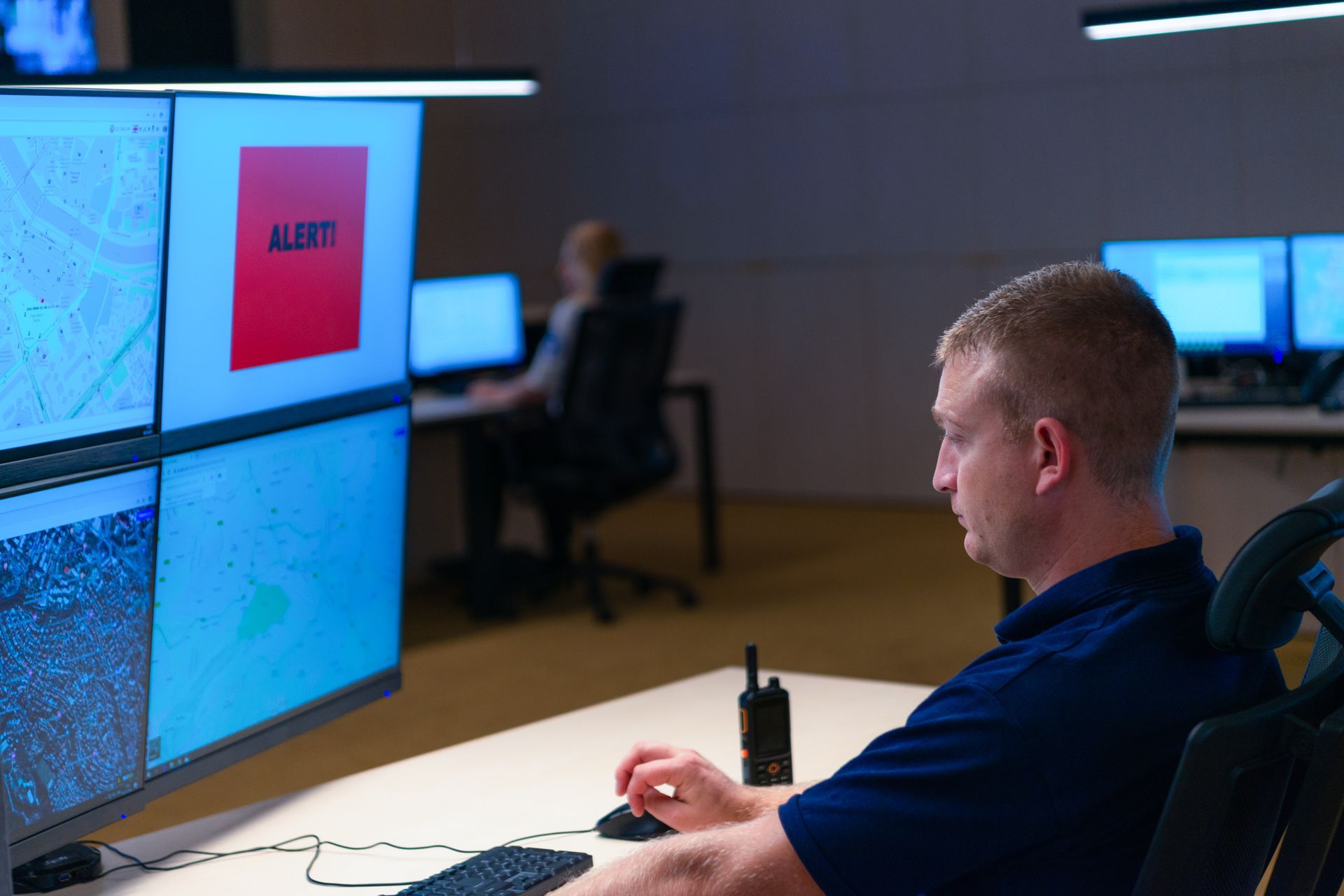 Man at desk monitors screens displaying maps, one with a red ALERT box, in a dark control room.