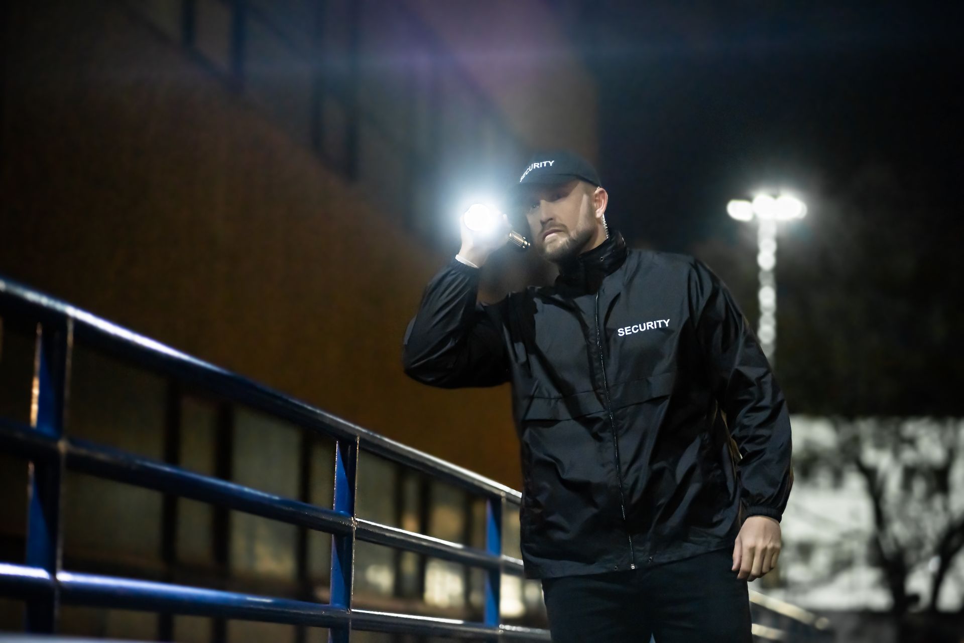 Security guard in black jacket, using a flashlight at night, near a building.