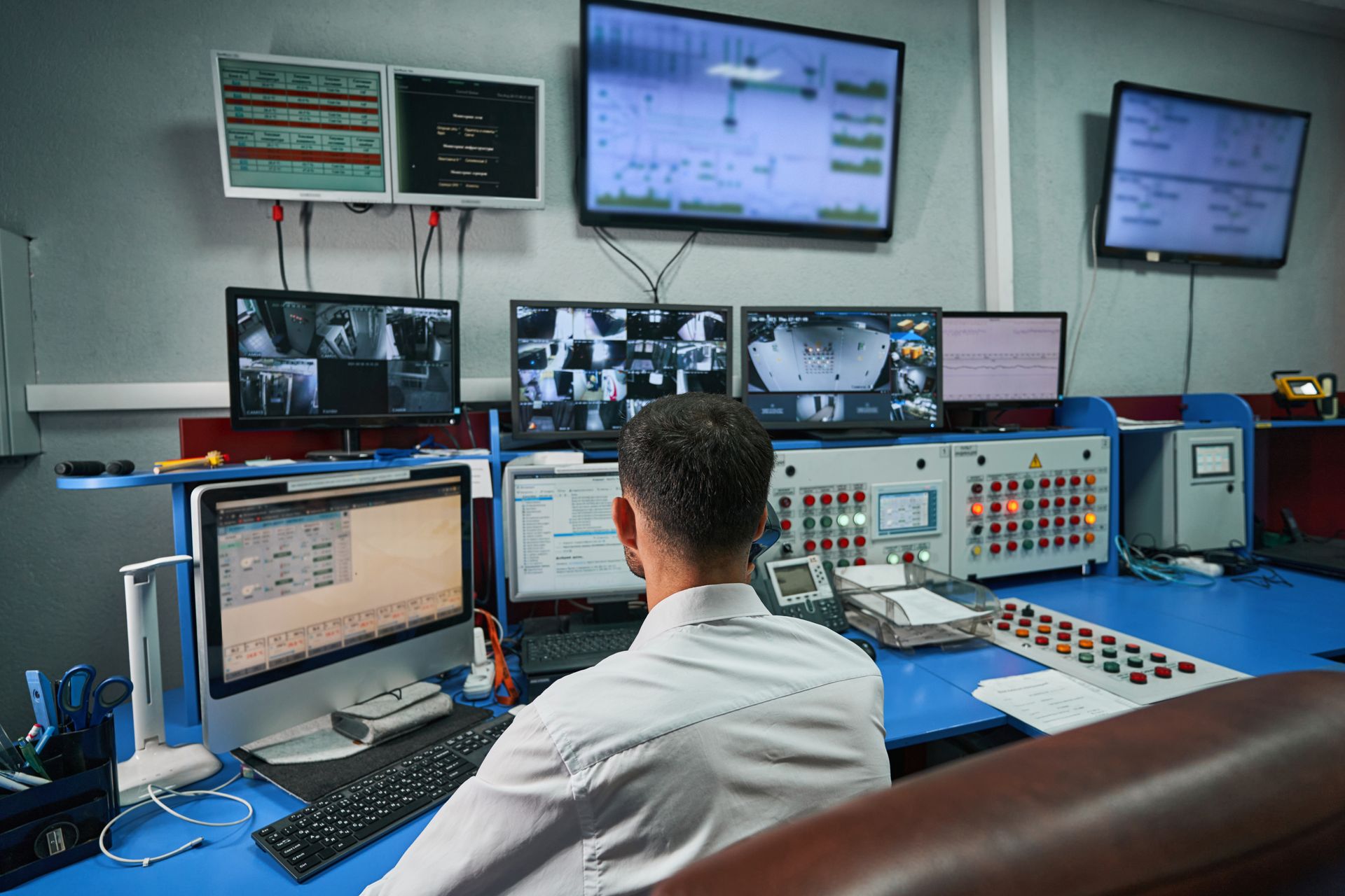 Man at a control panel with multiple monitors displaying data and camera feeds.