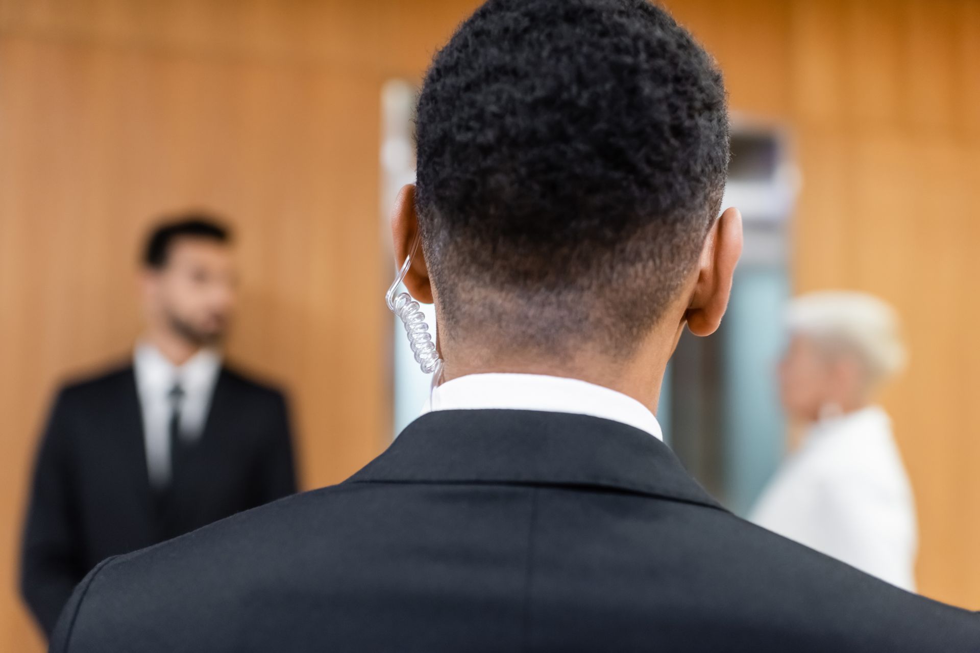 Back view of a person in a suit with earpiece, standing in front of two blurred figures in an interior space.