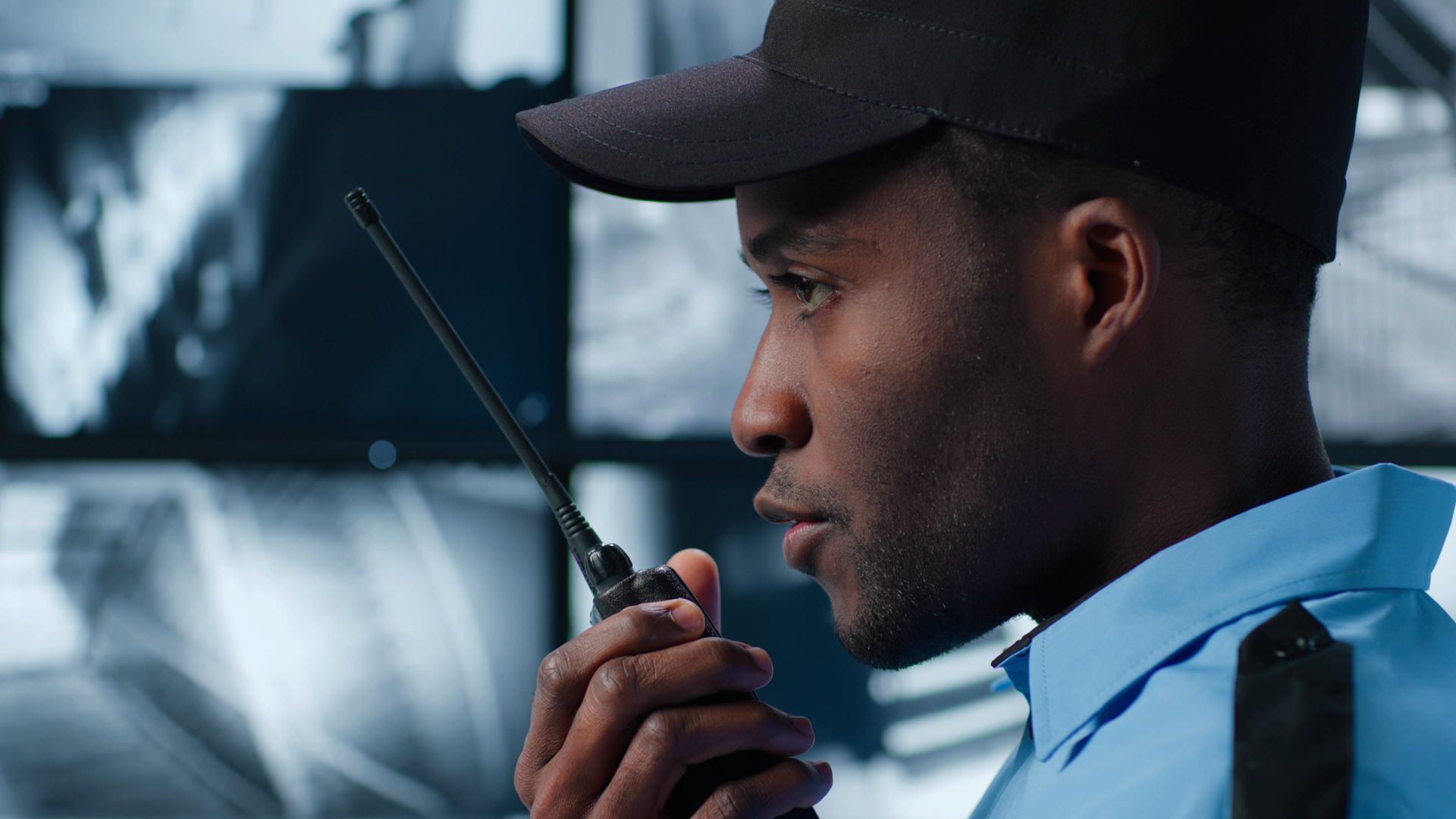 Security guard in uniform holds a walkie-talkie, looking focused in front of security monitors.