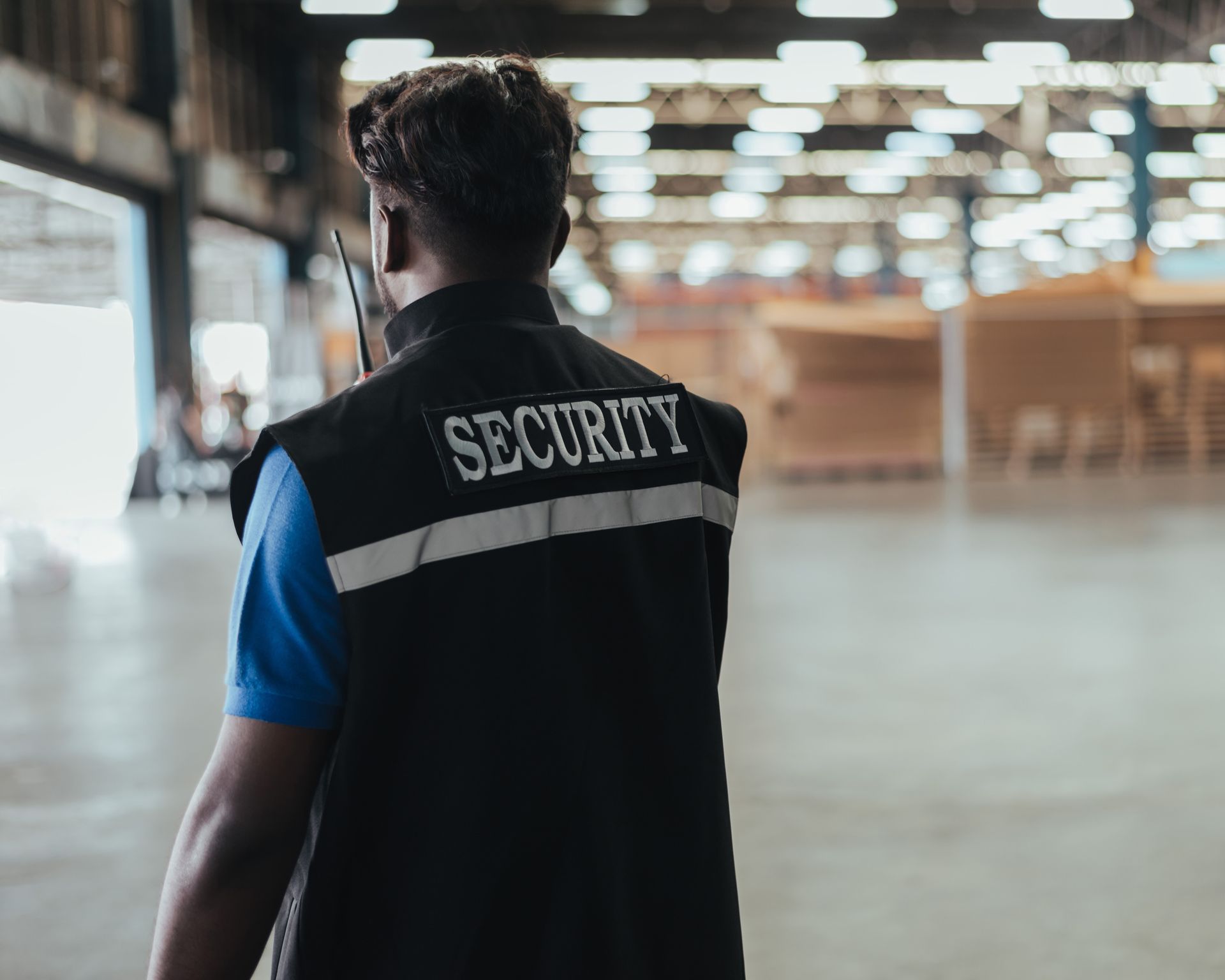 Security guard in black vest, back to the camera, standing in a warehouse, holding a radio.