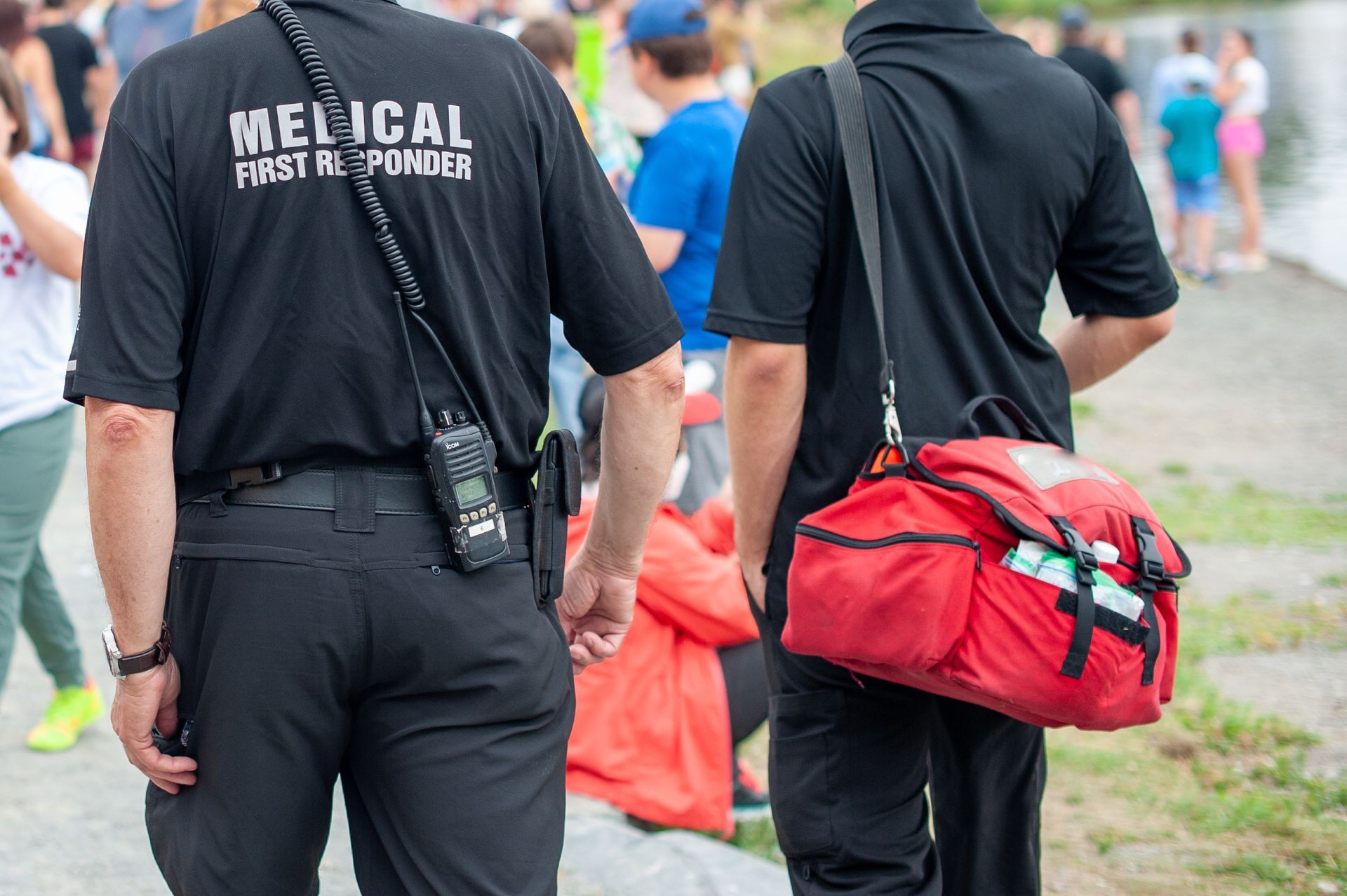 Two medical first responders in black uniforms; one with a radio, the other with a red medical bag, near a body of water.