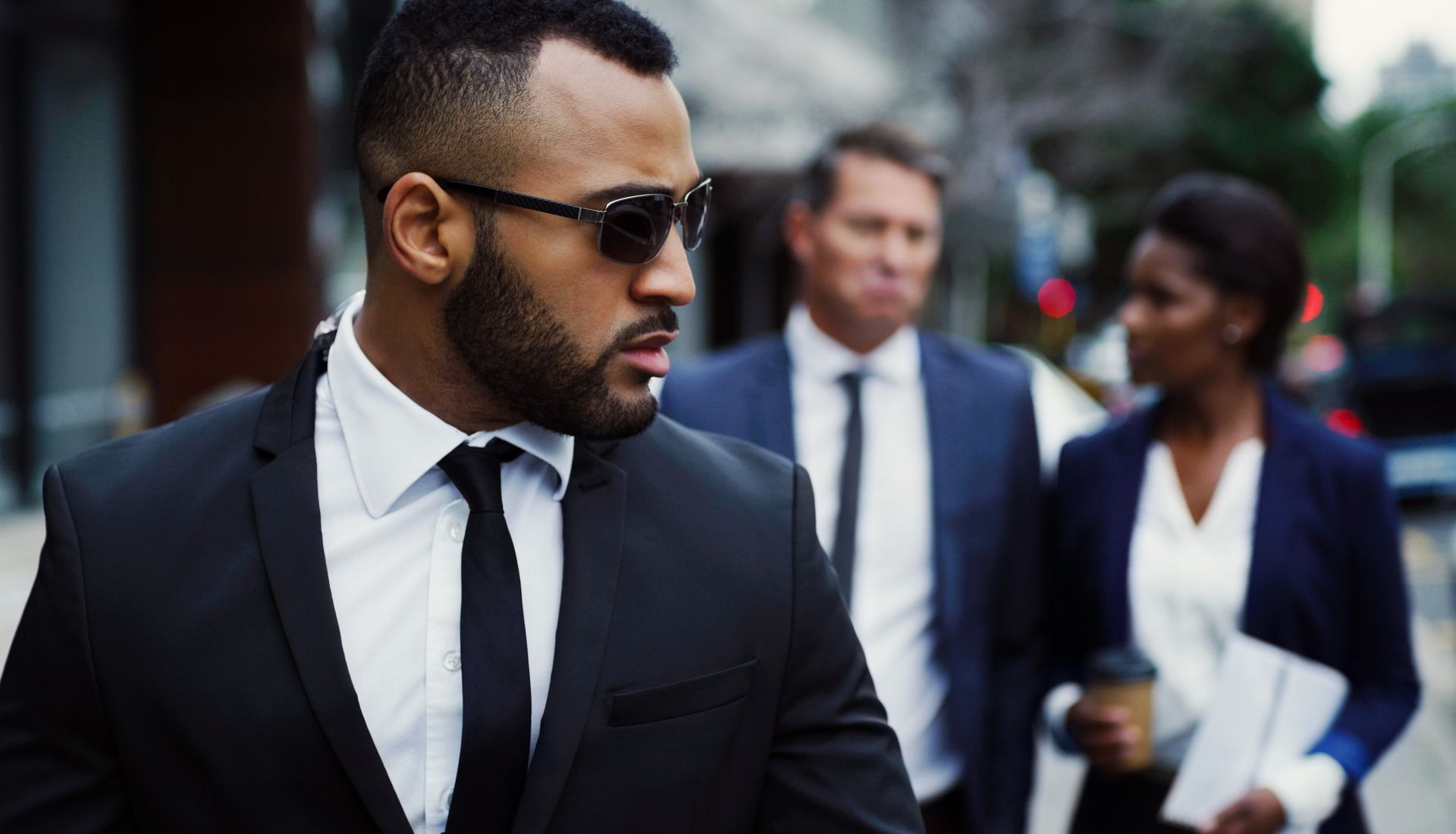 Man in sunglasses, black suit, looks back while walking with two people in business attire on a city street.
