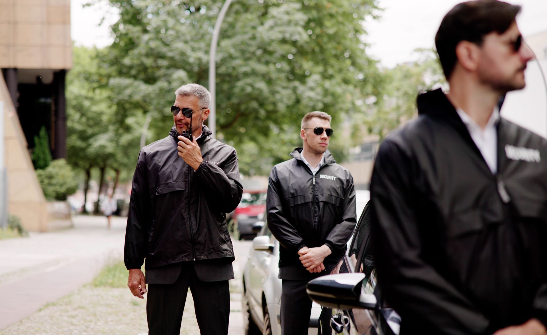 Three security guards in black uniforms standing near a parked car; one uses a walkie-talkie.