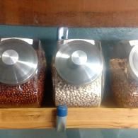 Three glass jars of beans on a wooden shelf with metal lids and a blue spout in front