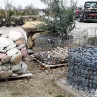 Wire cages filled with rocks and sandbags outdoors in a muddy yard, with a small tree in the center