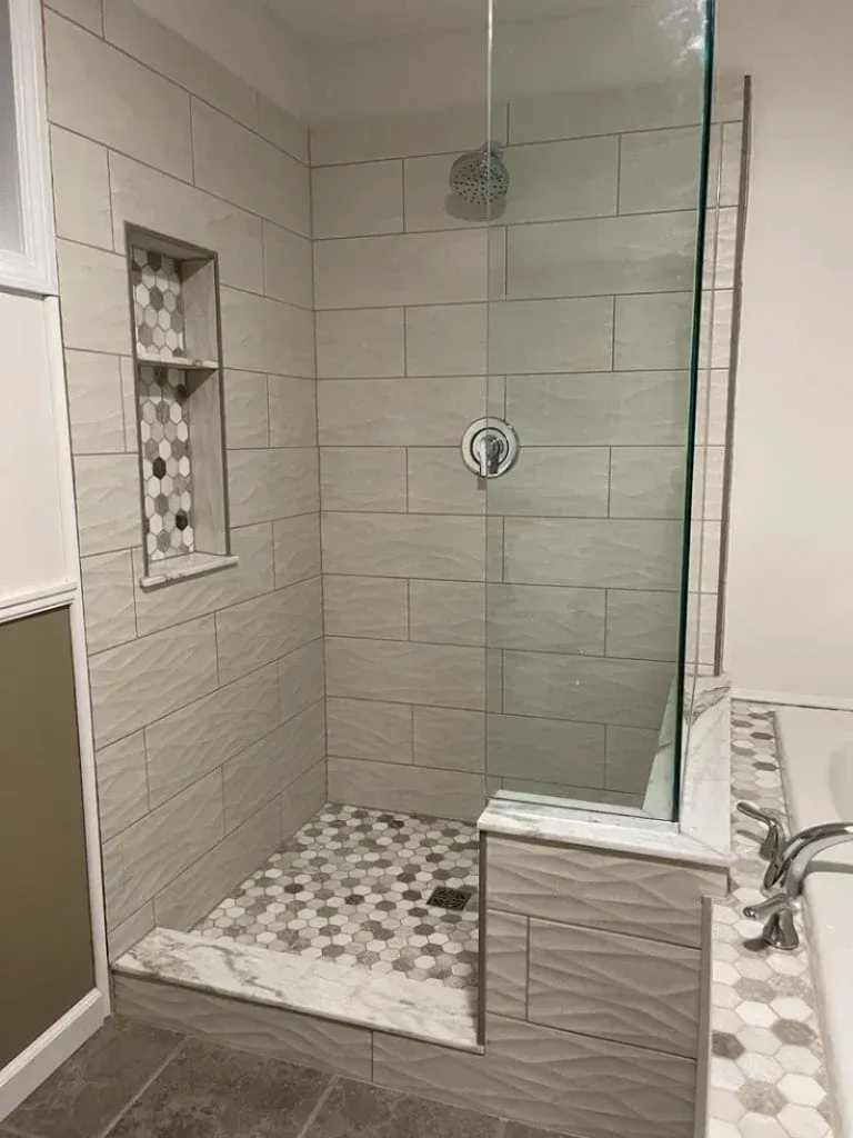Newly tiled shower with glass door and built-in shelf; light gray and hexagonal tiles.