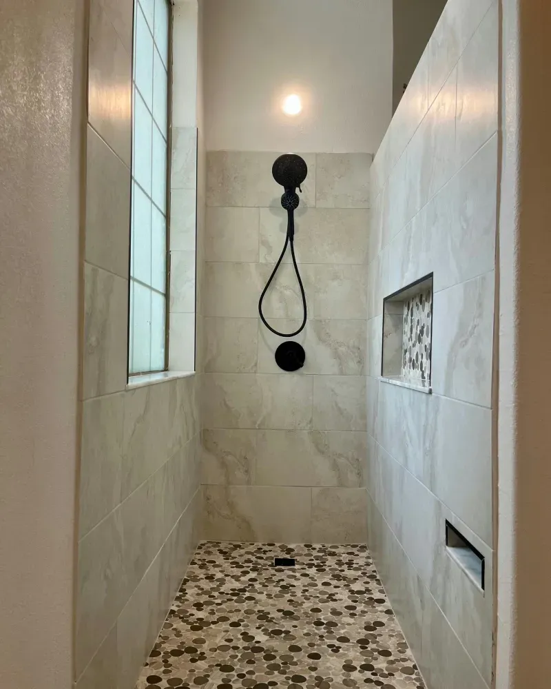 Shower with neutral-toned tiled walls, pebble floor, black showerhead, and a window.